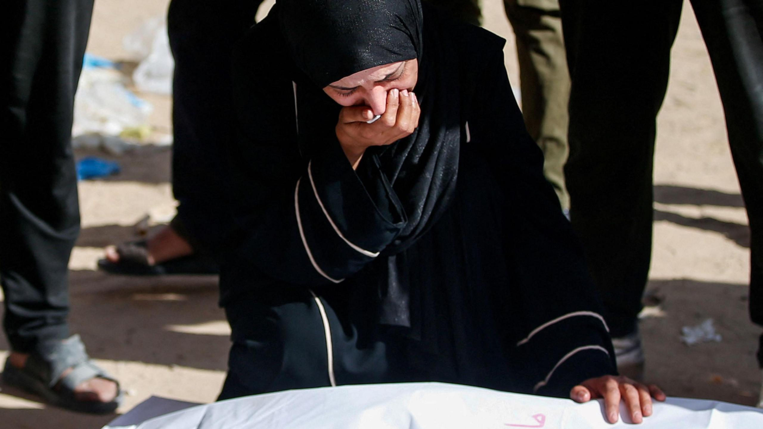 A crying woman wearing dark clothing and a hijab kneels, one hand to her mouth, the other on the shrouded body of a corpse