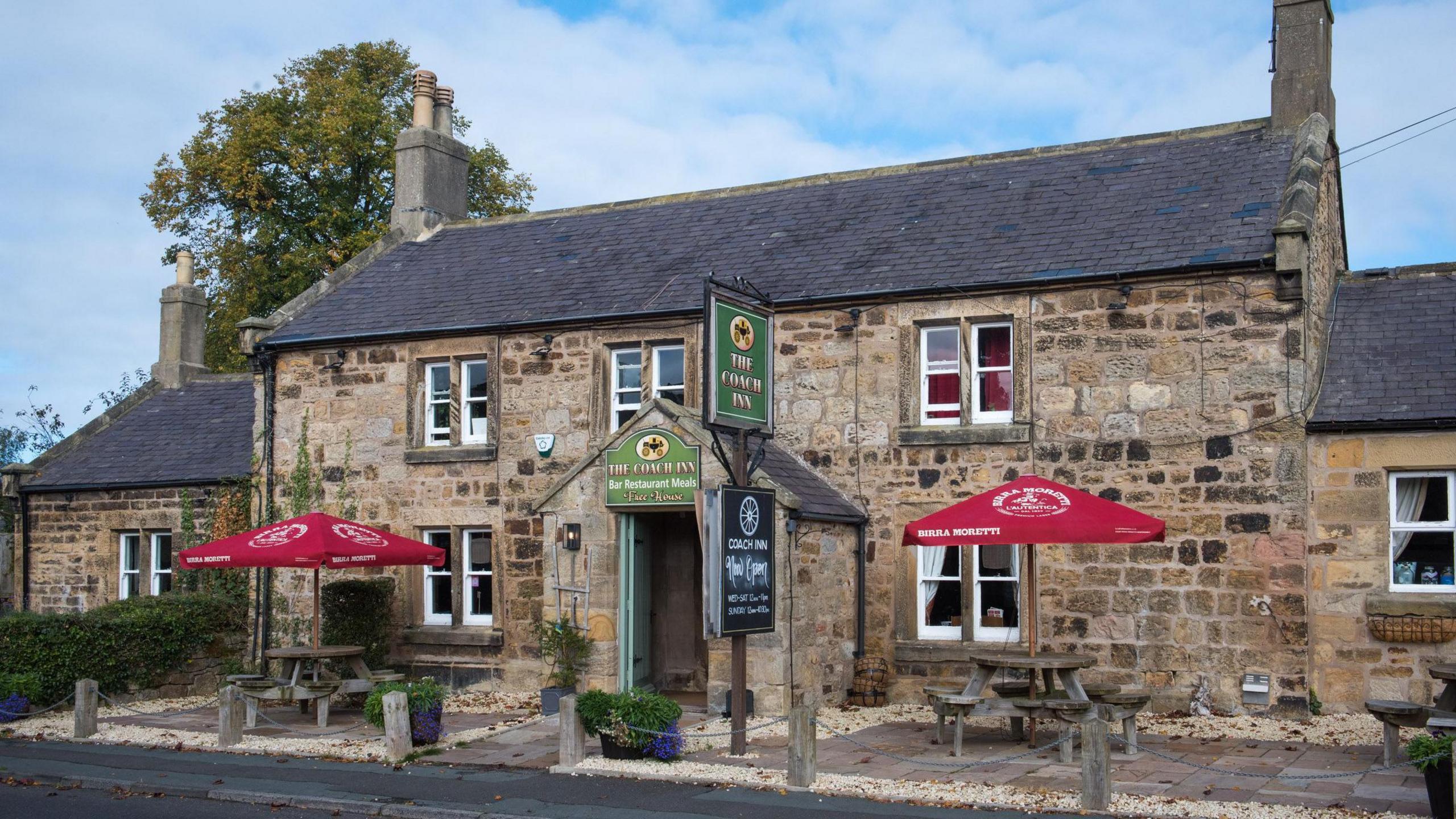 A pub from the outside with tables and red umbrellas. It is an old stone building with a chimney at each end and a sign saying Coach Inn outside and over the door