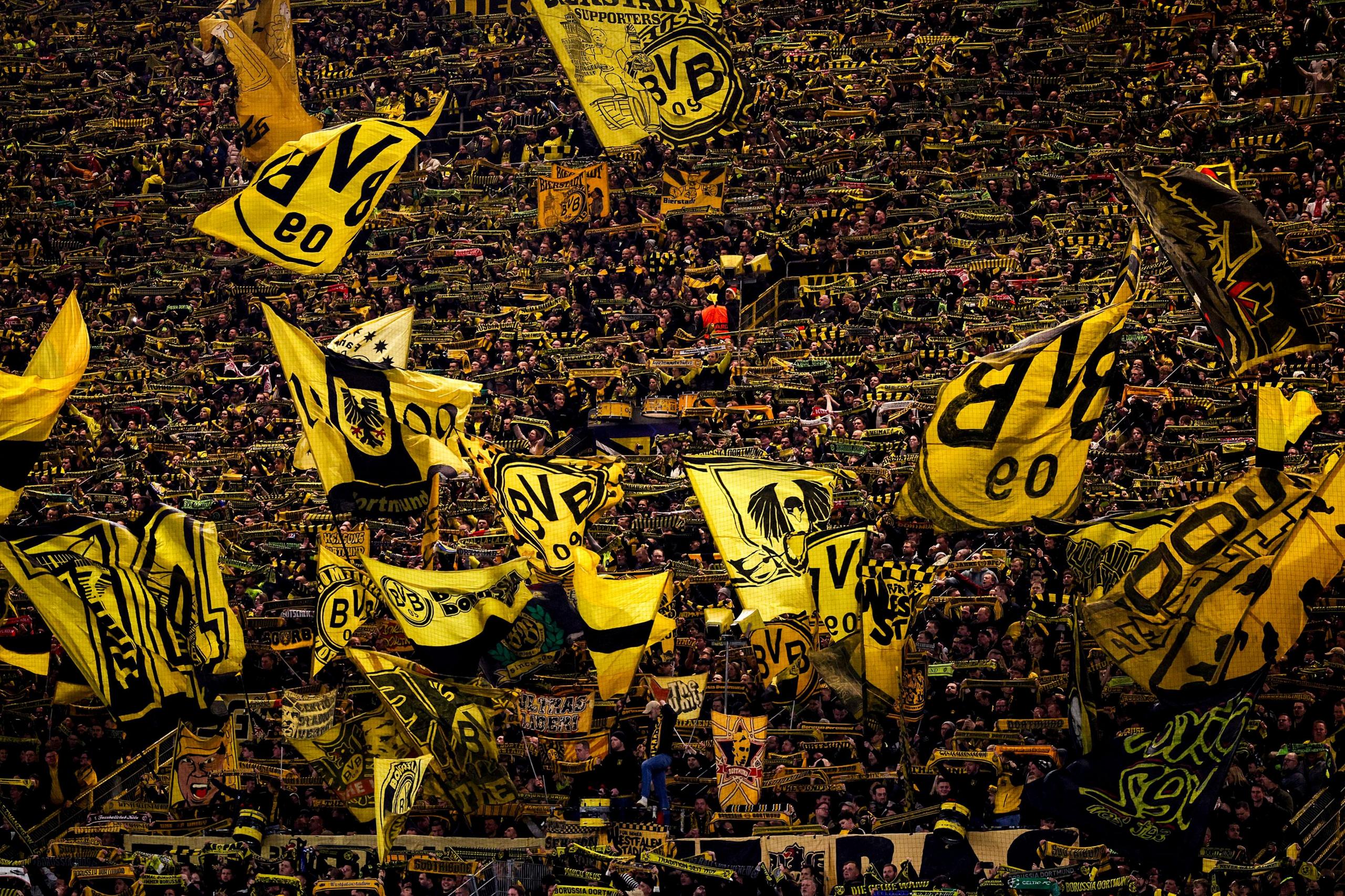 Una densa multitud de seguidores del Borussia Dortmund llena las gradas del estadio, ondeando grandes banderas, bufandas y pancartas amarillas y negras, creando un mar vibrante de colores del equipo.