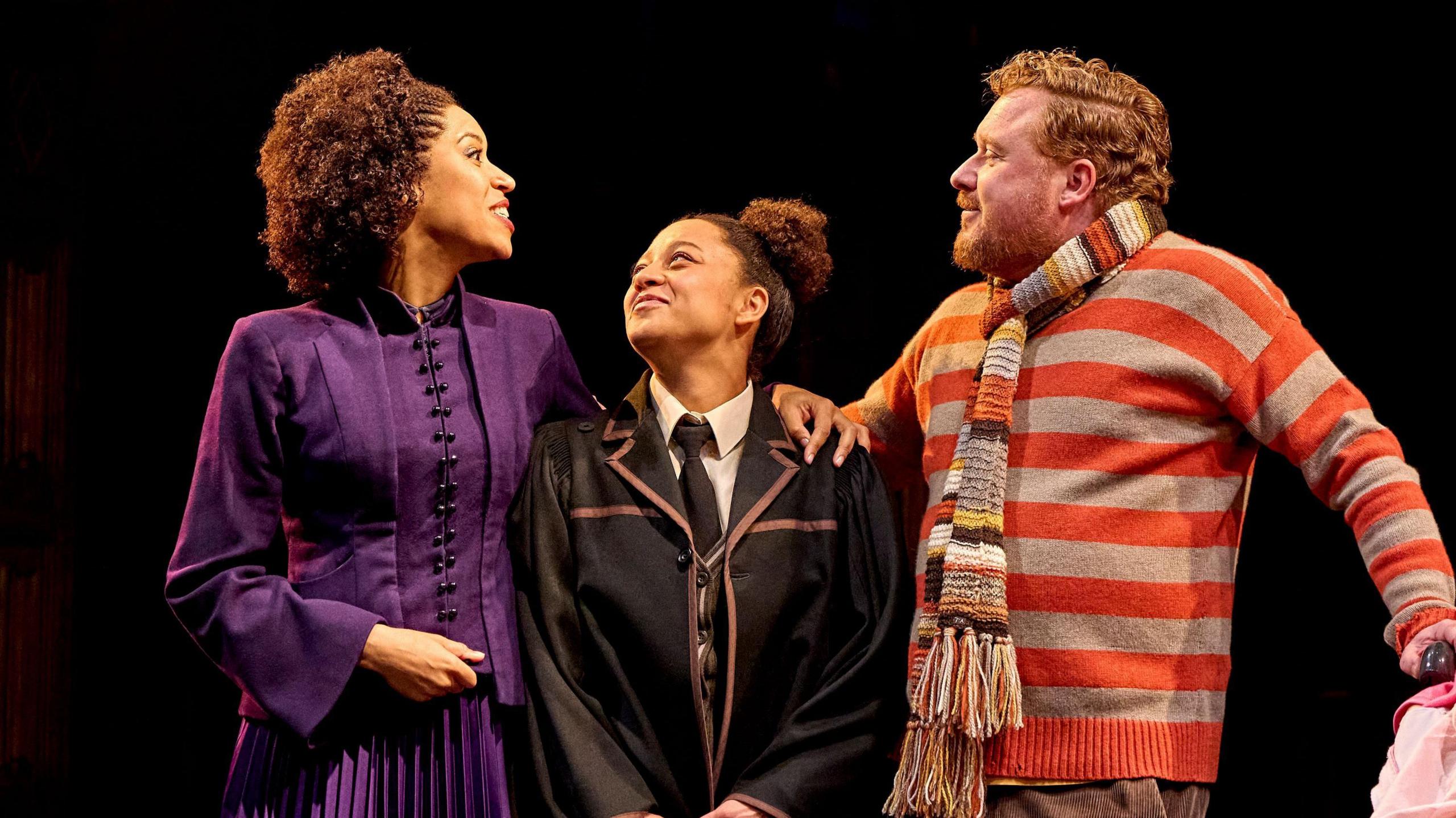 Three people on stage. There is a man and a woman and in the middle there is a teenager.
The woman is woman is wearing a purple gown and the teenager is in her uniform and is smiling at her mum.
The man is wearing a orange and grey striped jumper and a striped woolen scarf