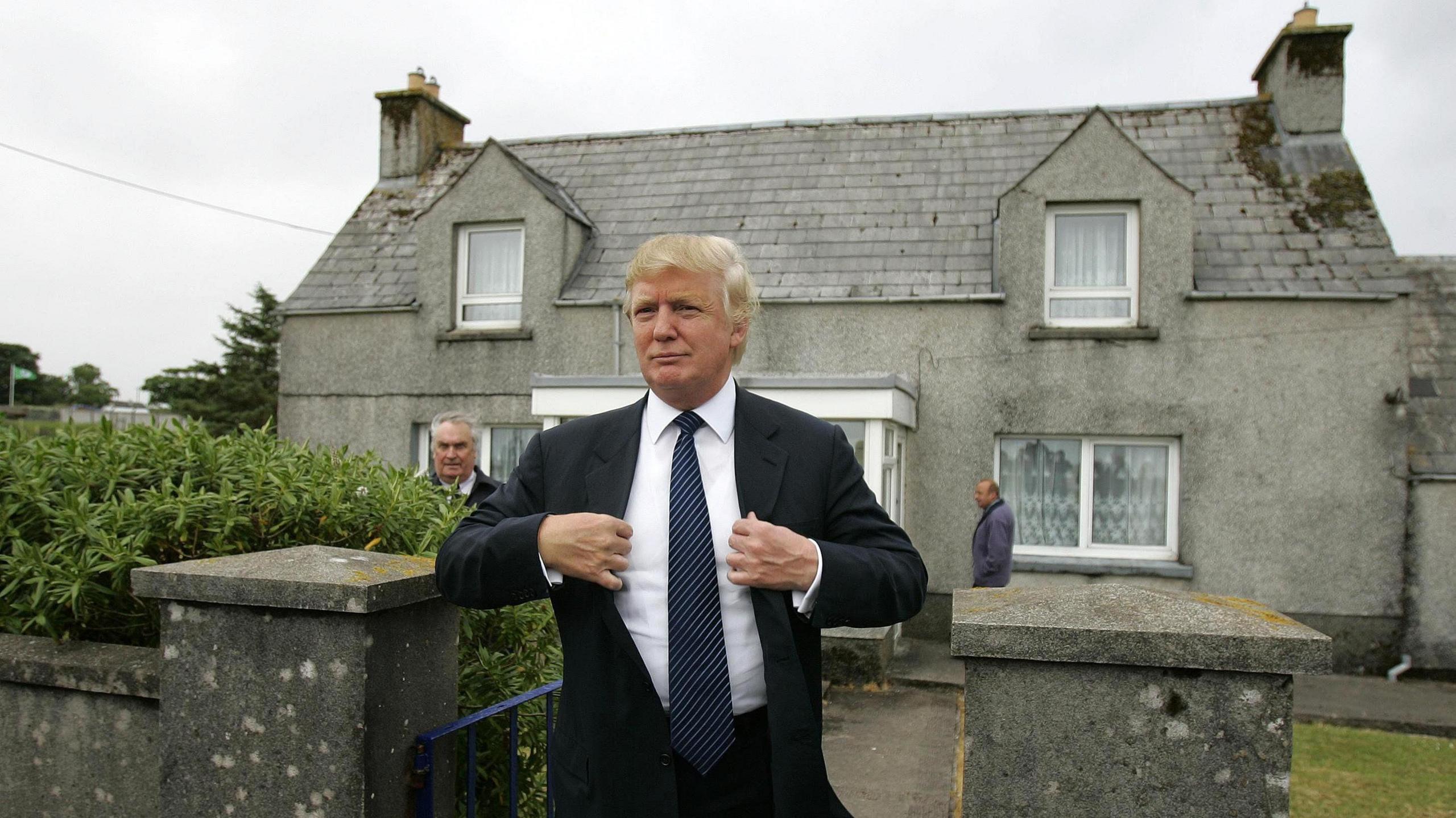 Donald Trump in dark blue suit, plain shite shirt and dark blue striped tie, standing outside a grey-harled house on a grey day. He is smiling and appears to be fixing his lapels.