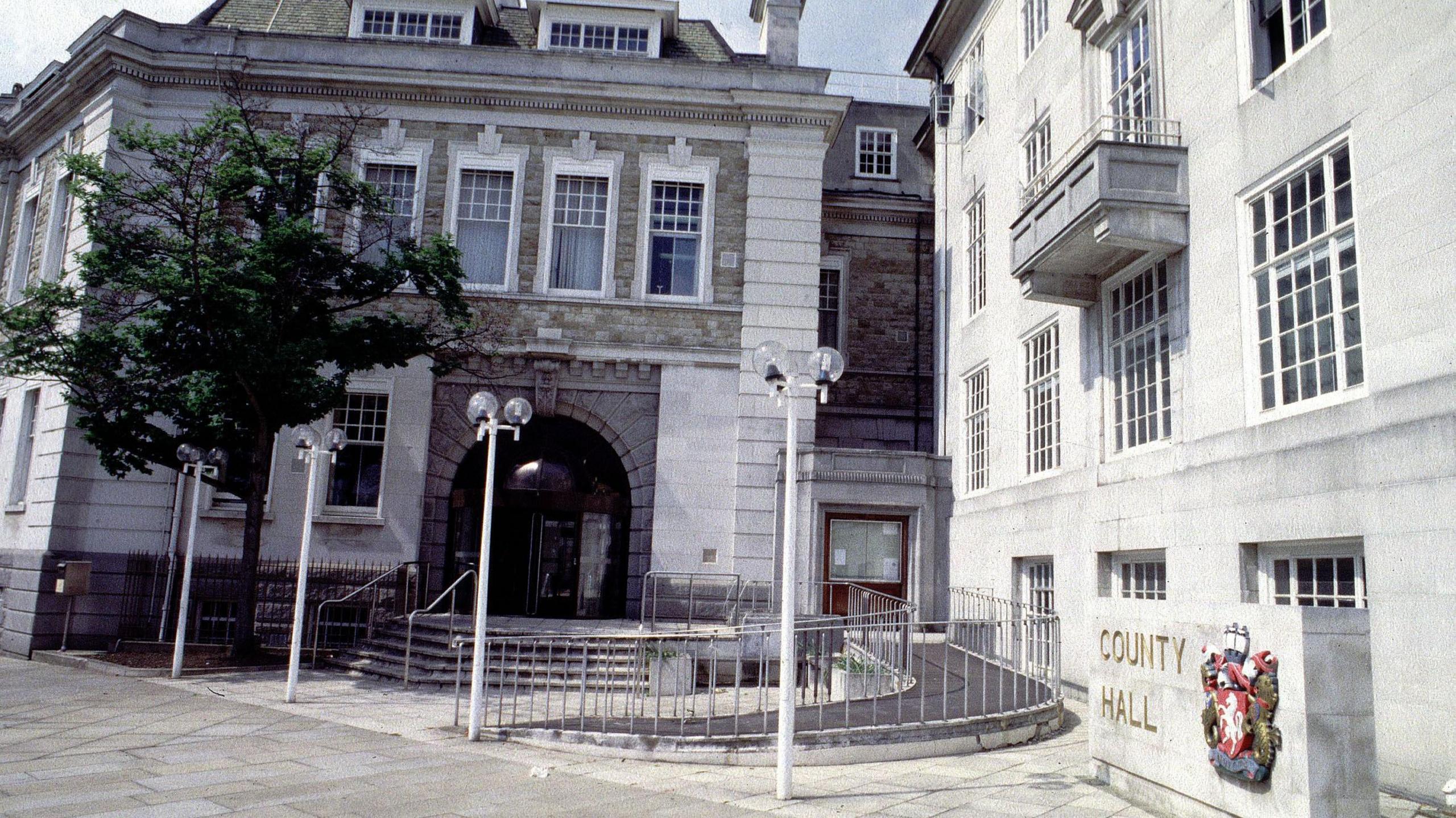 An exterior view of County Hall in Maidstone, a building of white portland stone with a dark glass fronted door and four lampposts in front of it.