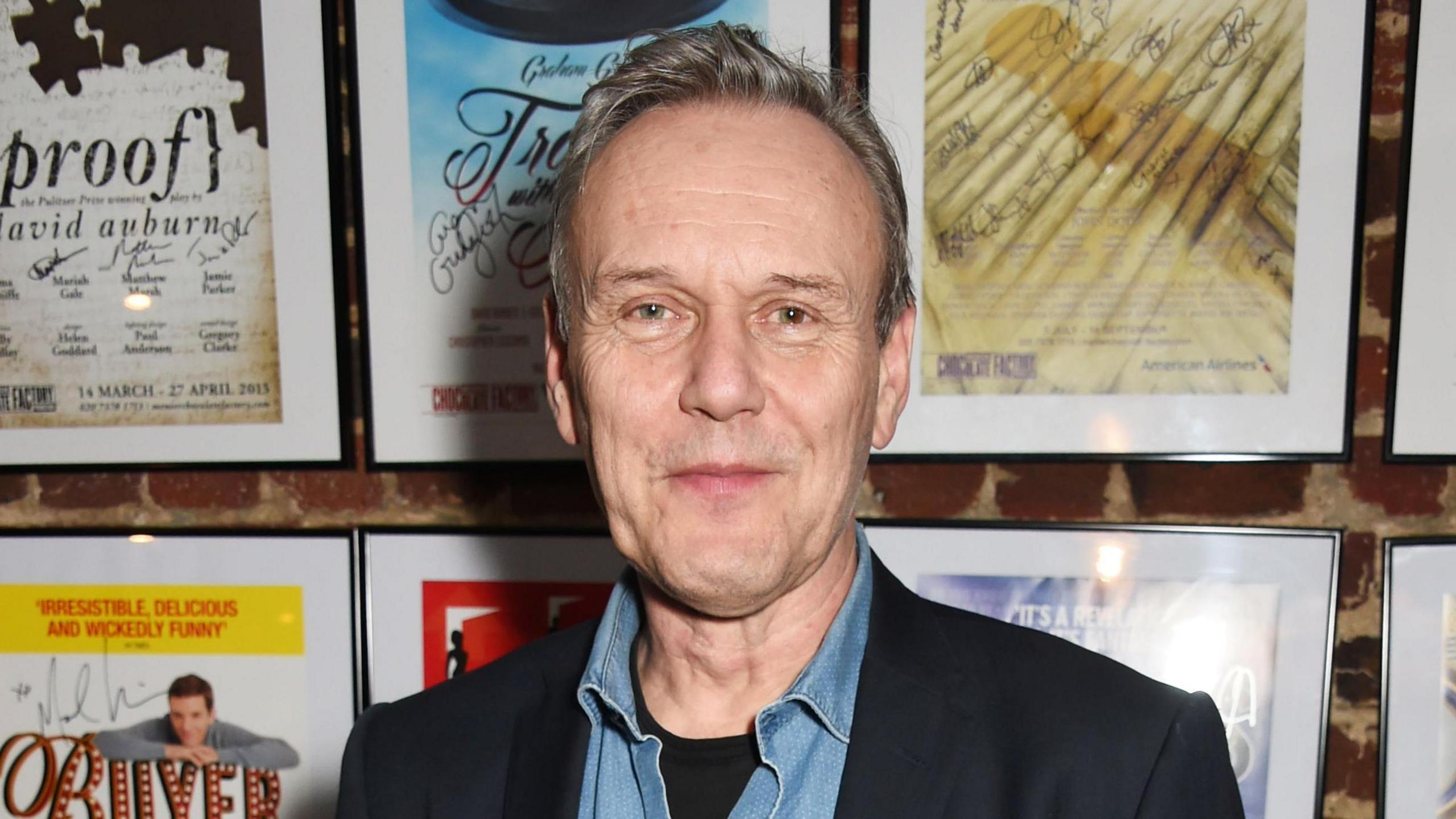 Anthony Head is looking towards the camera smiling. He has short grey and brown hair and is wearing a black blazer with a denim shirt underneath. He is standing in front of a wall which has small posted displayed in frames. 