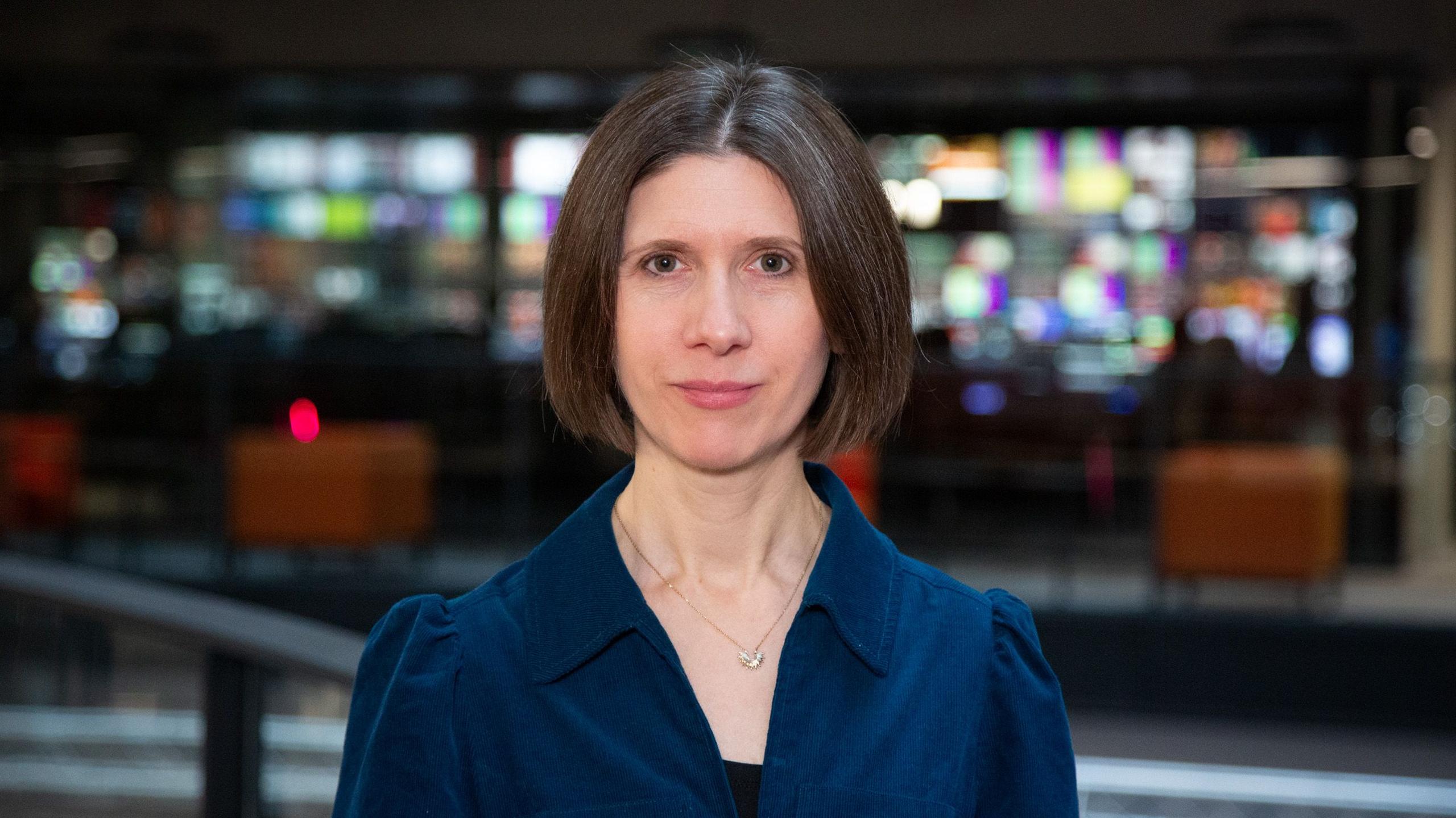 Haf Elgar o Awyr Iach Cymru yn edrych yn syth at y camera. Mae ganddi wallt brown wedi ei torri mewn bob, ac yn gwisgo ffrog glas. Mae hi'n mewn adeilad gyda fodern gyda seddi a sgrins teledu yn y pellter tu ol 