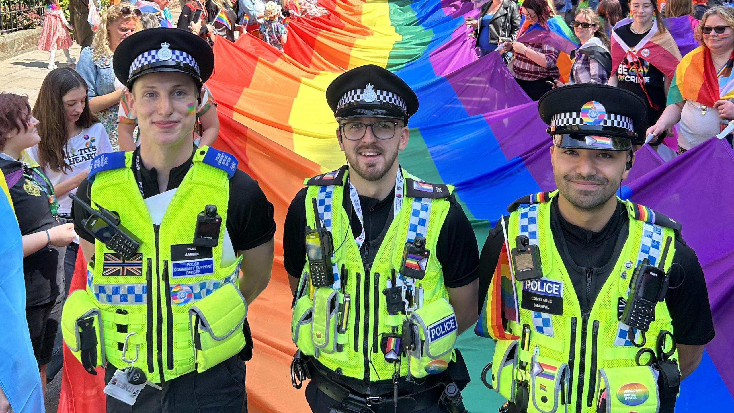 Three police officers stand smiling in front of a large pride flag held by members of the public at York Pride 2023. They are in uniform, with pride flag stickers and patches adorning their vests. One officer has a pride flag painted onto her cheek.