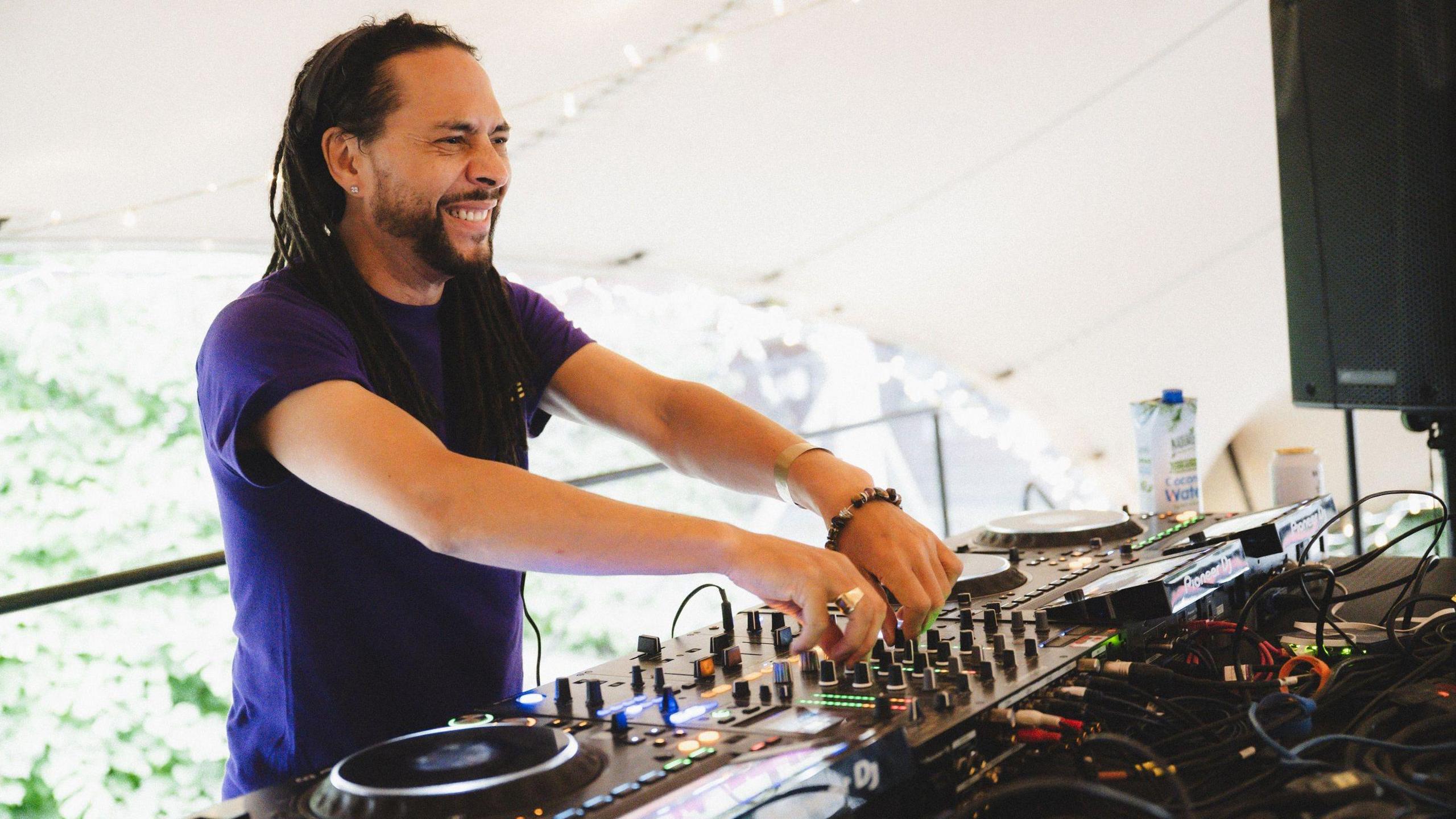 DJ Roni Size smiles as he has both hands on the mixer of some DJ decks. He is wearing a purple T-shirt and has long black dreadlocks.