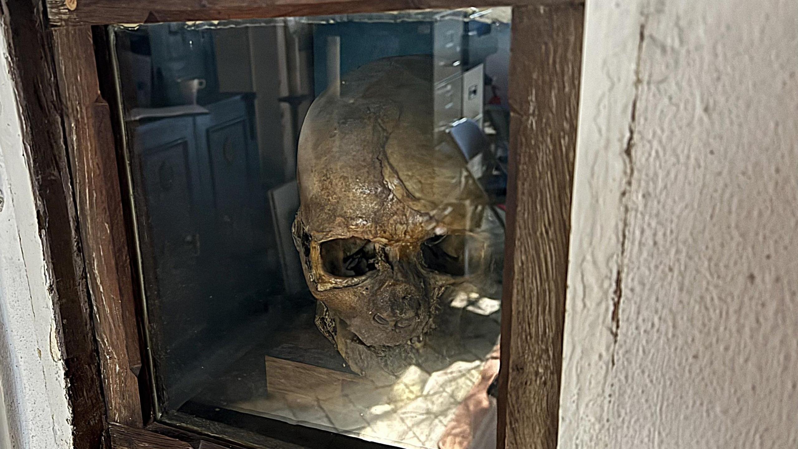 A mummified skull sitting on a small wooden block behind a glass screen.  