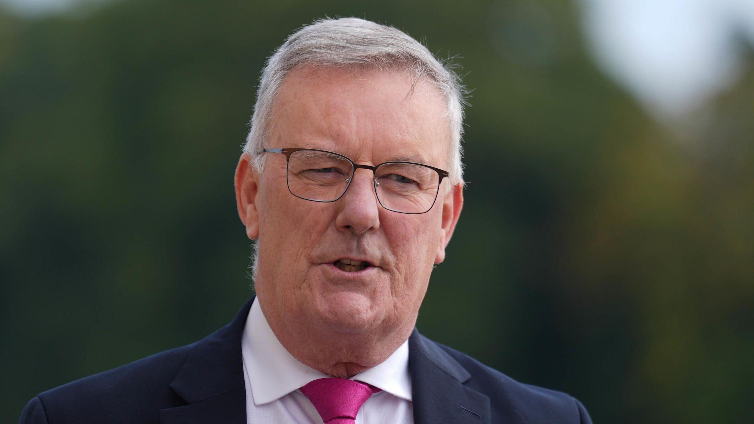 Mike Nesbitt speaking outside, he has short white/grey hair and is wearing thin framed glasses. He is also wearing a navy suit, a white shirt and a hot pink tie.