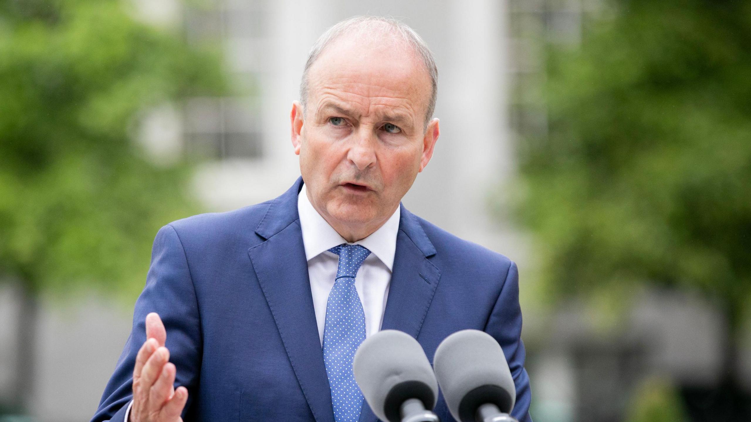 Mr Martin wearing a navy blue suit, blue spotty tie and white shirt as he talks into two mics.