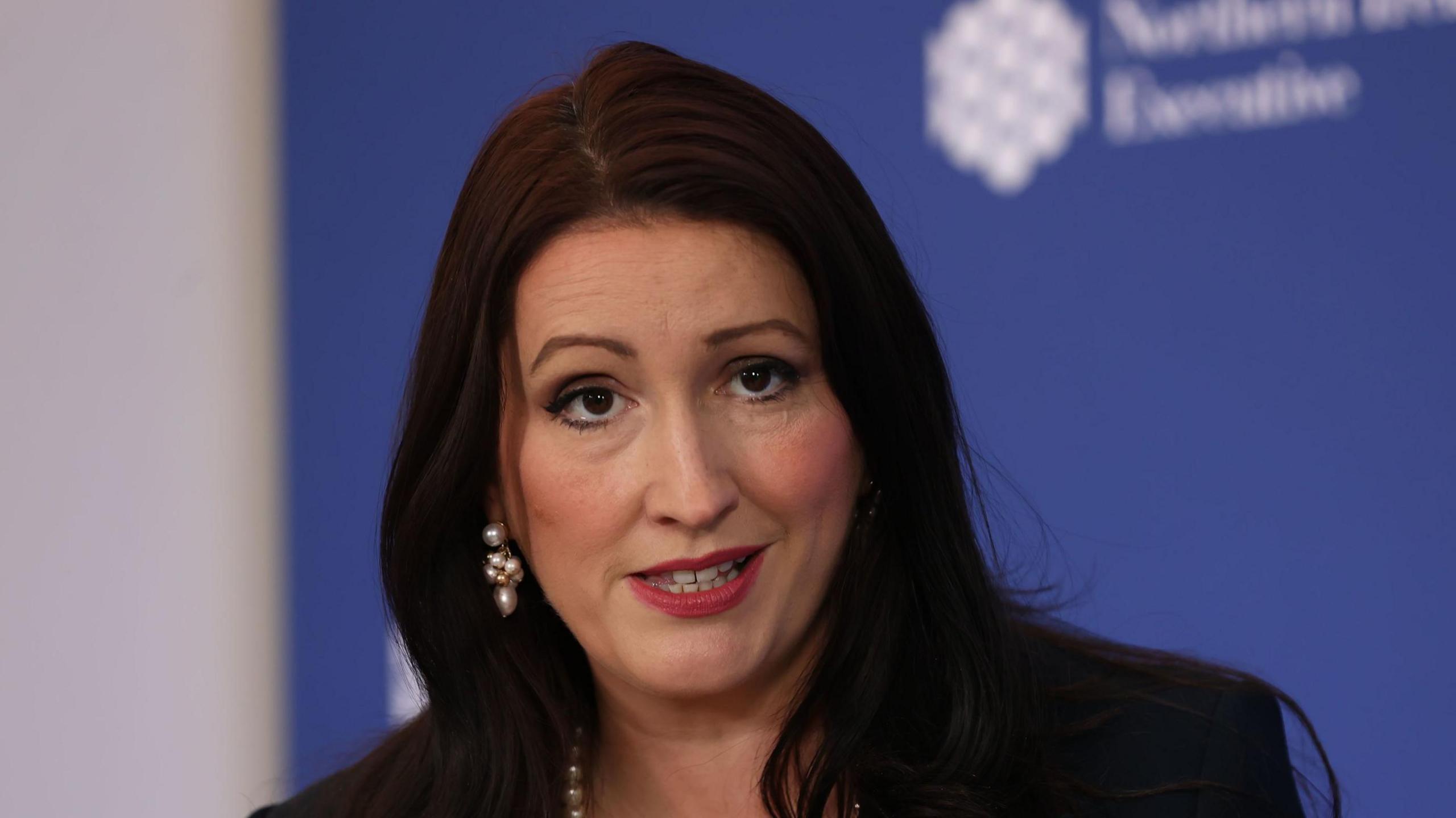 Emma Little-Pengelly has long black hair and is wearing makeup, including pink lipstick. She has a pearl necklace and earrings and is wearing a black blazer.