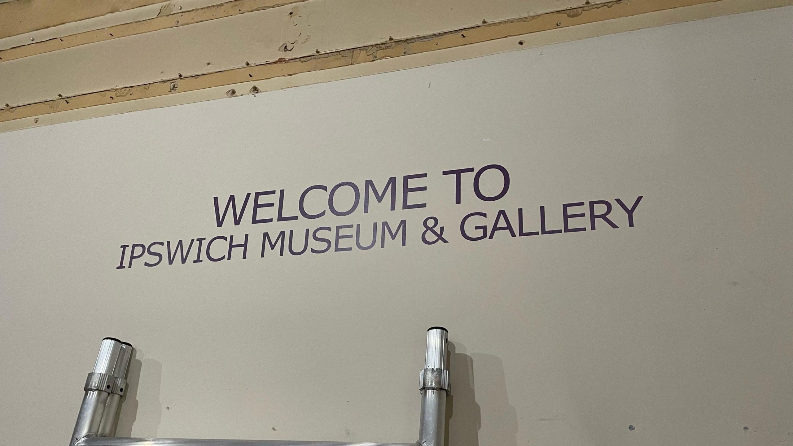 A sign is pictured which reads, 'Welcome to Ipswich Museum & Gallery'. The writing is in a black font on a white background. The top of a ladder can be seen resting underneath.