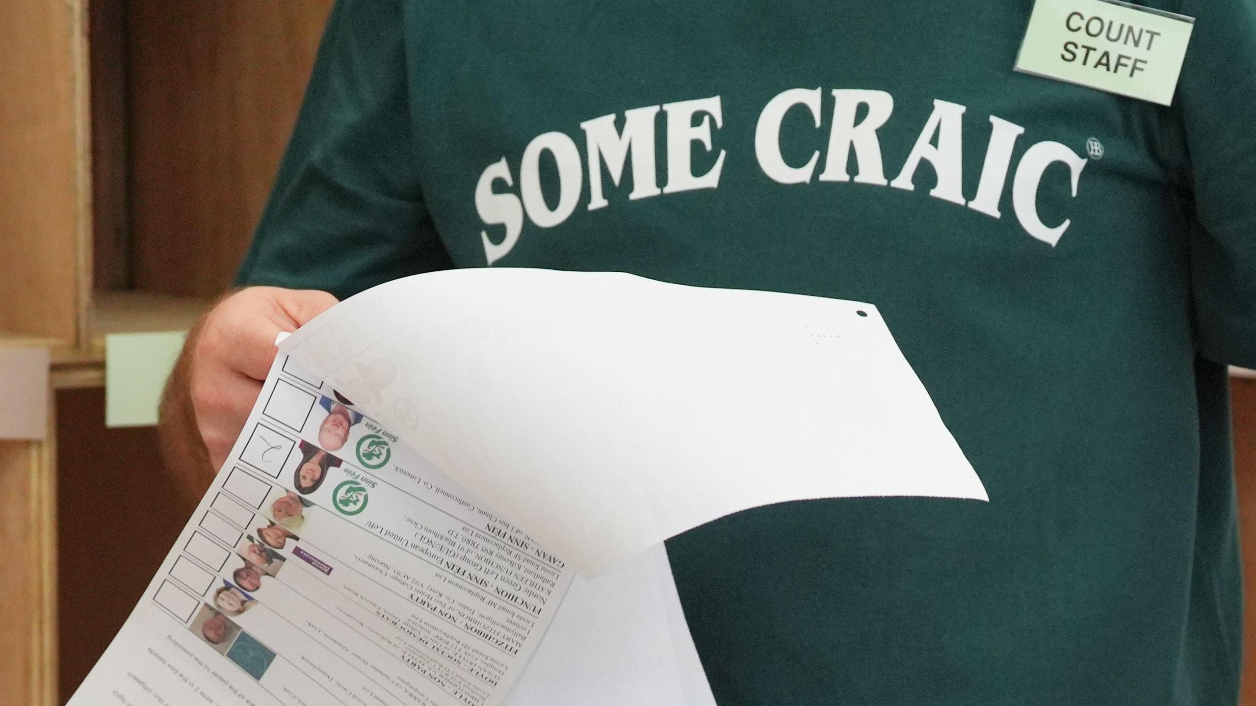 A counter is holding some election ballots in his hand. He's wearing a green jumper that says some craic and a badge that says, count staff.