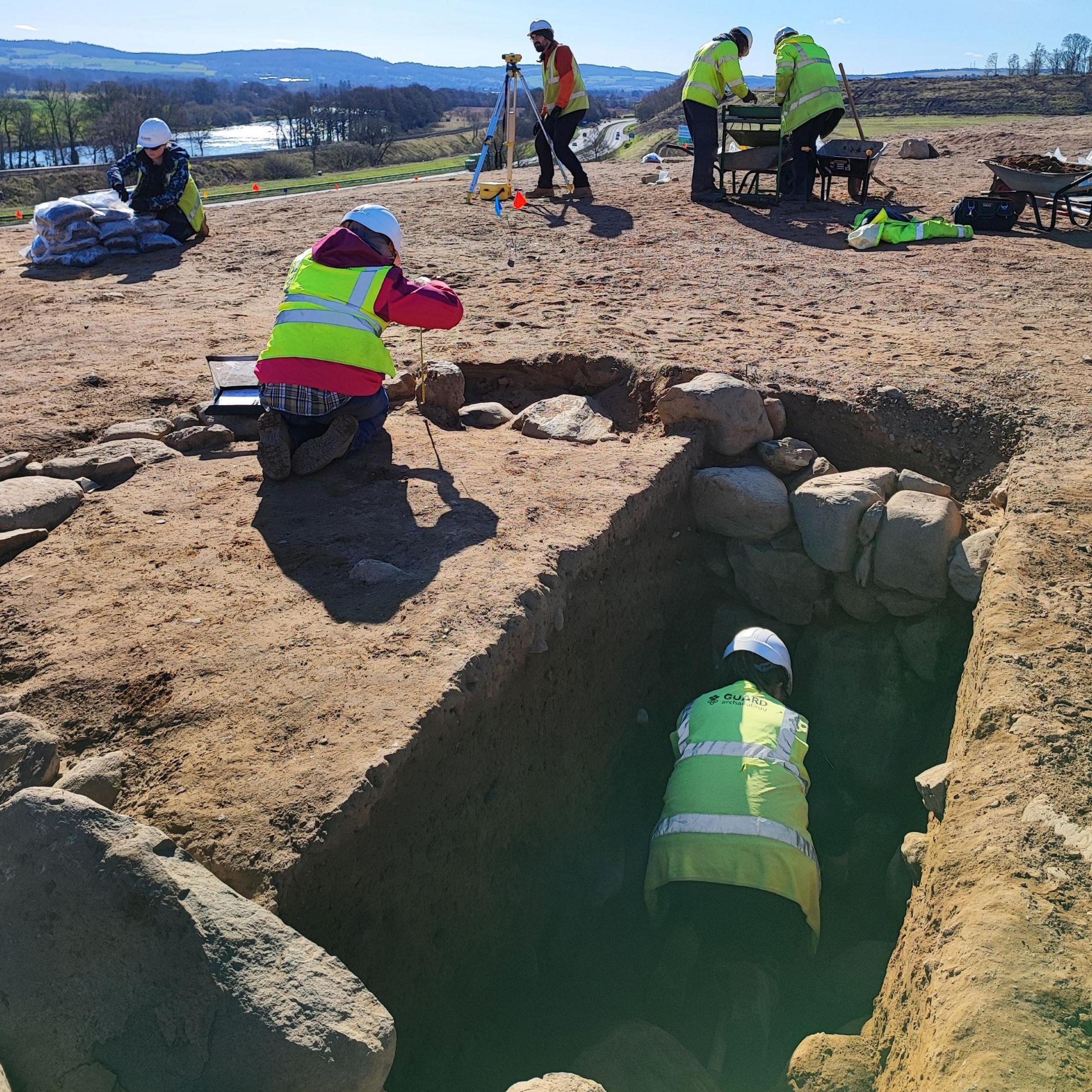 Guard Archaeologists excavating Broxy Kennels Fort. The archaeologists are working in and around deep trenches. They are wearing white hard hats and high visibility jackets.
