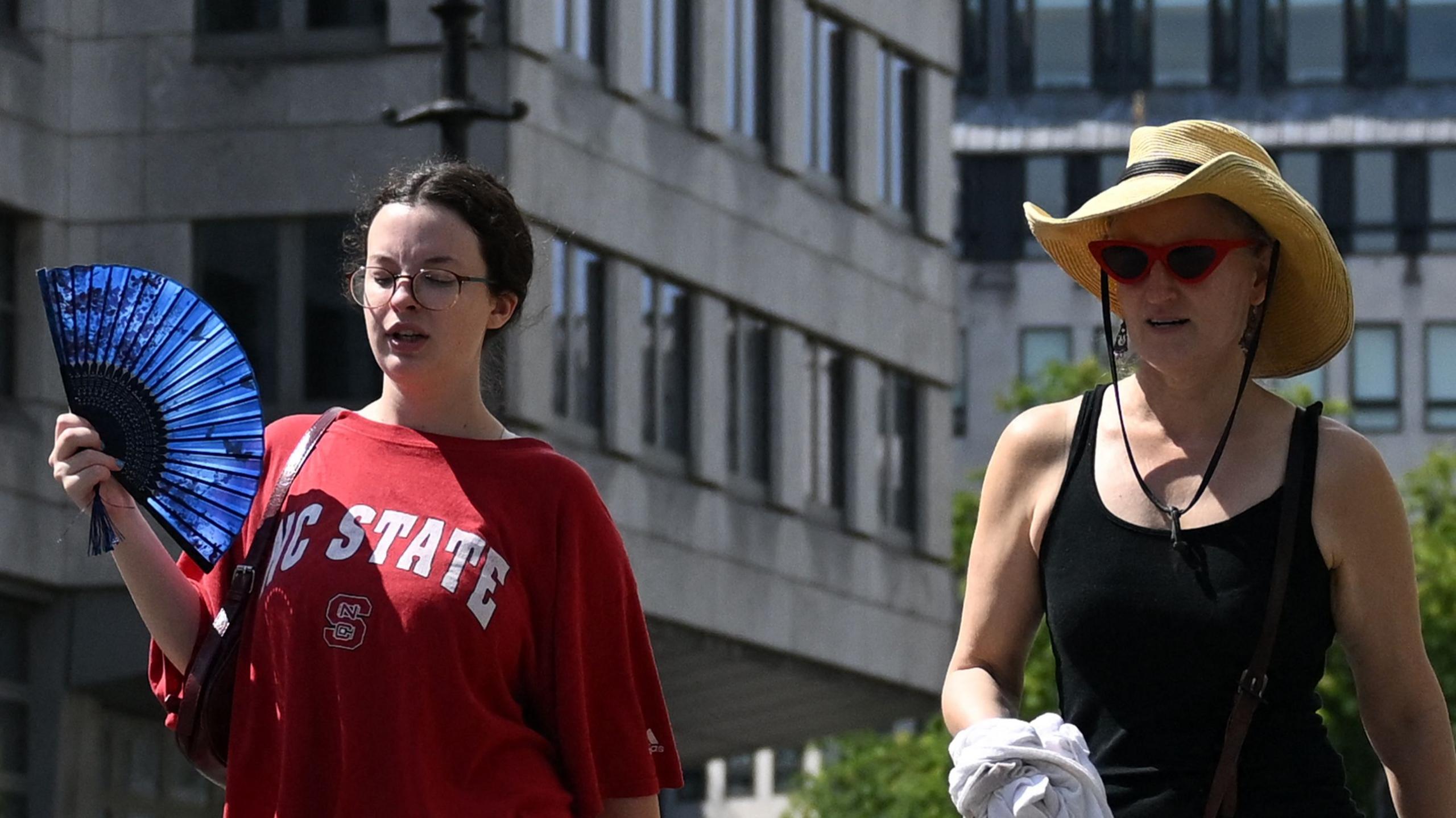 Two women walking, one uses a blue manual fan and the other wears a wide-brimmed straw hat in the sun. They look warm, and the sun beams on them.