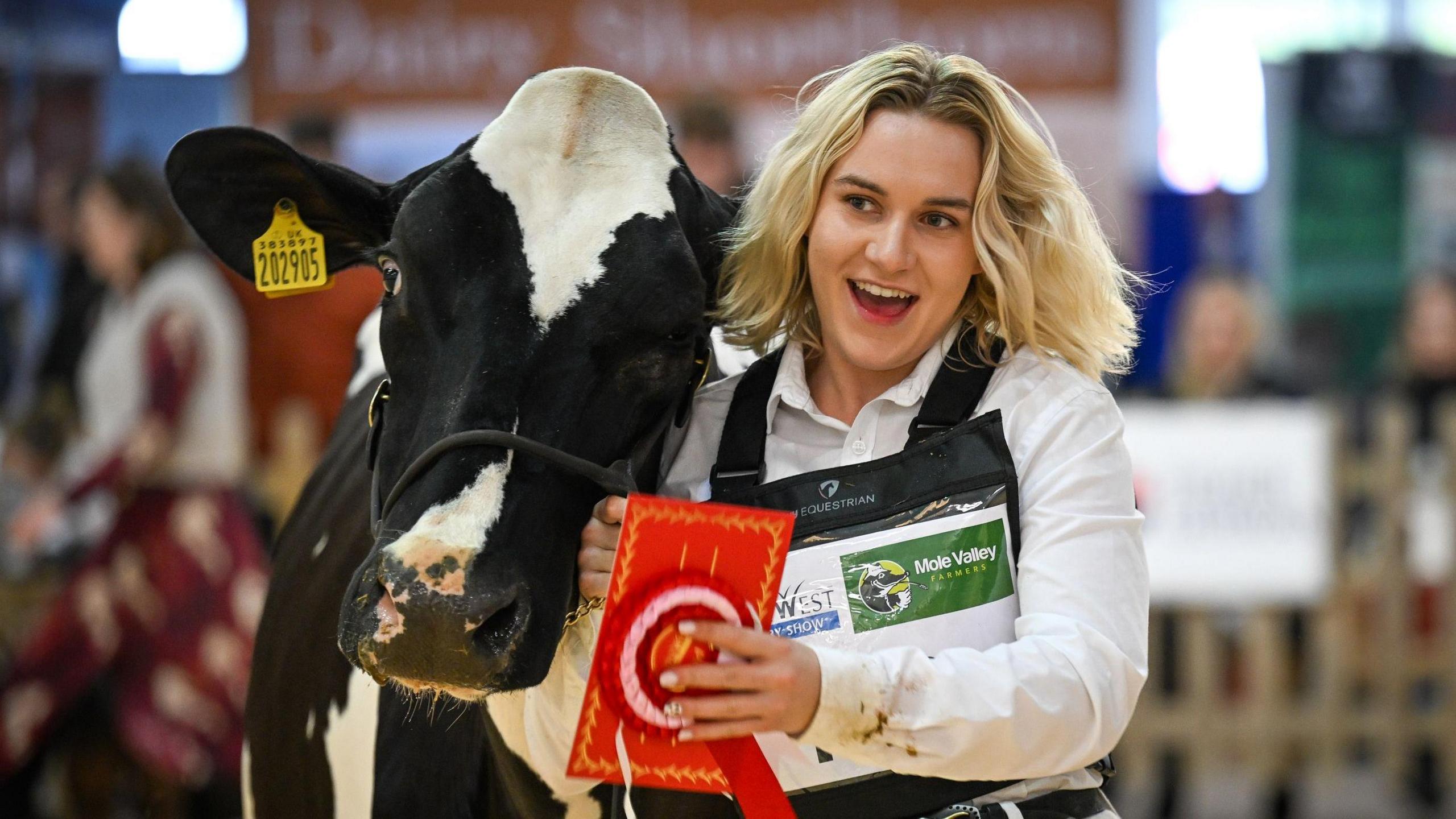 A woman is standing next to a calf. She has blonde hair and is holding a prize. 