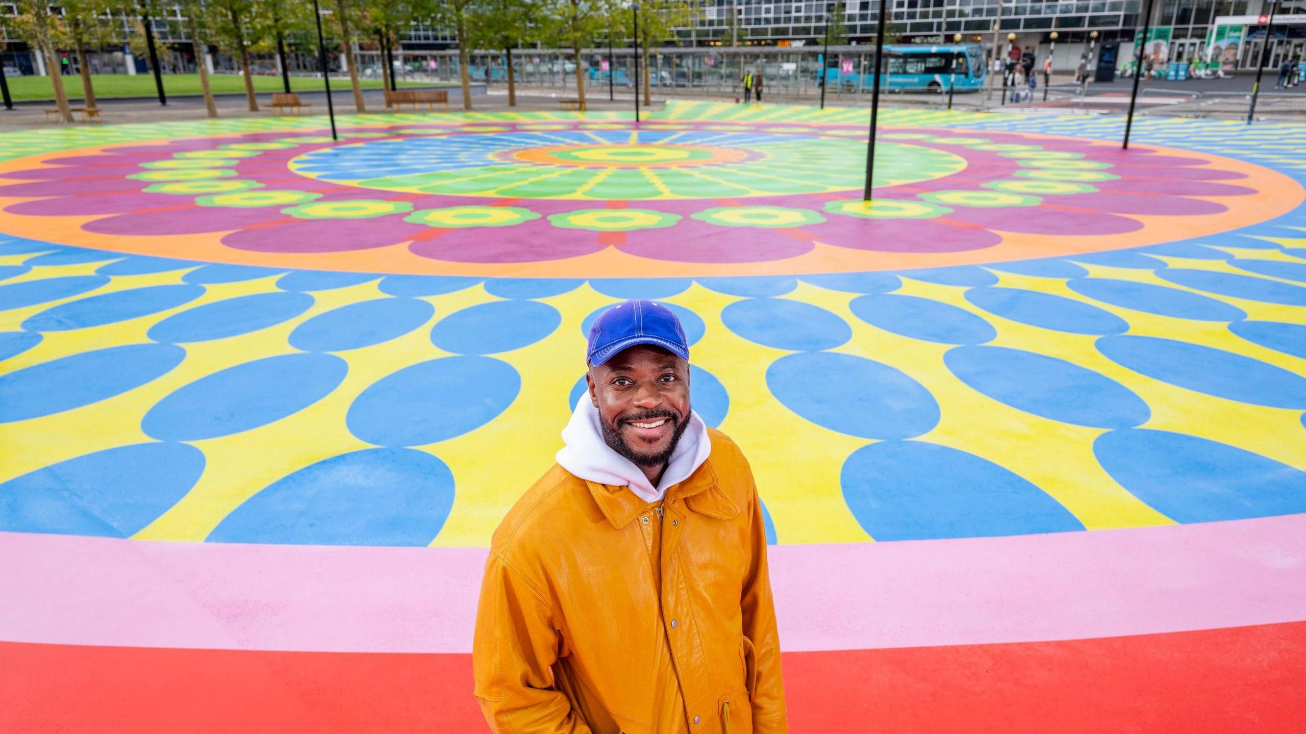 Yinka stands in front of the flower artwork, smiling at the camera. He wears an orange coat over a white hoody, and a blue peaked cap.