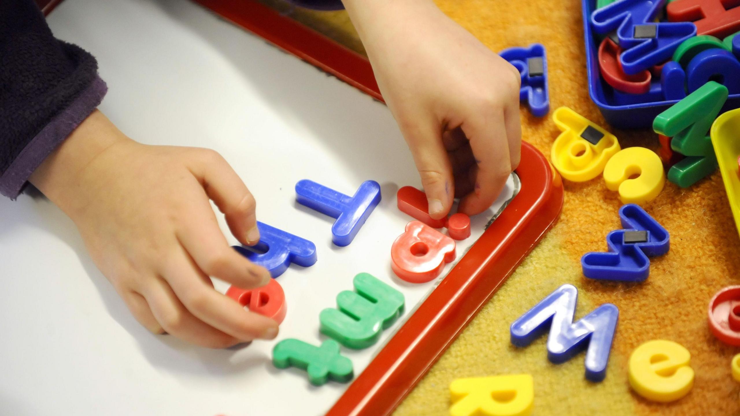 A child is seen playing with plastic letters with magnets attached on a magnetic white board. The board has a red frame.