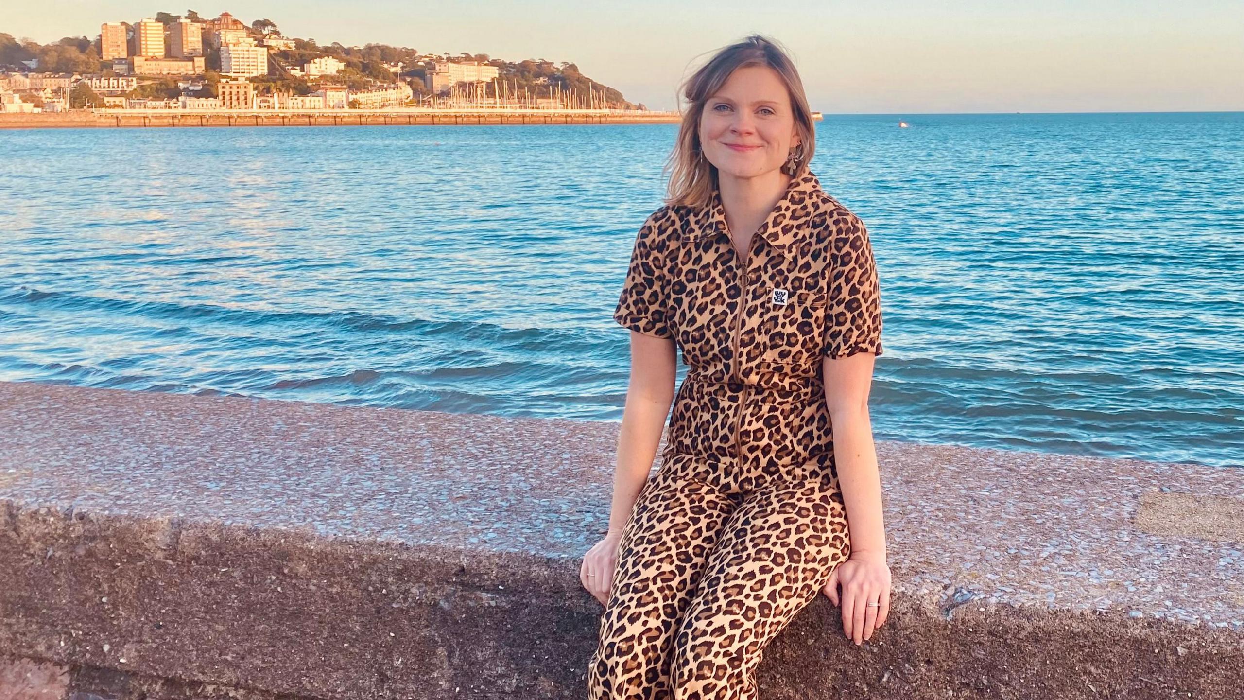 Laura wearing a leopard print jumpsuit sat on a wall by the sea with a peninsula of land in the background