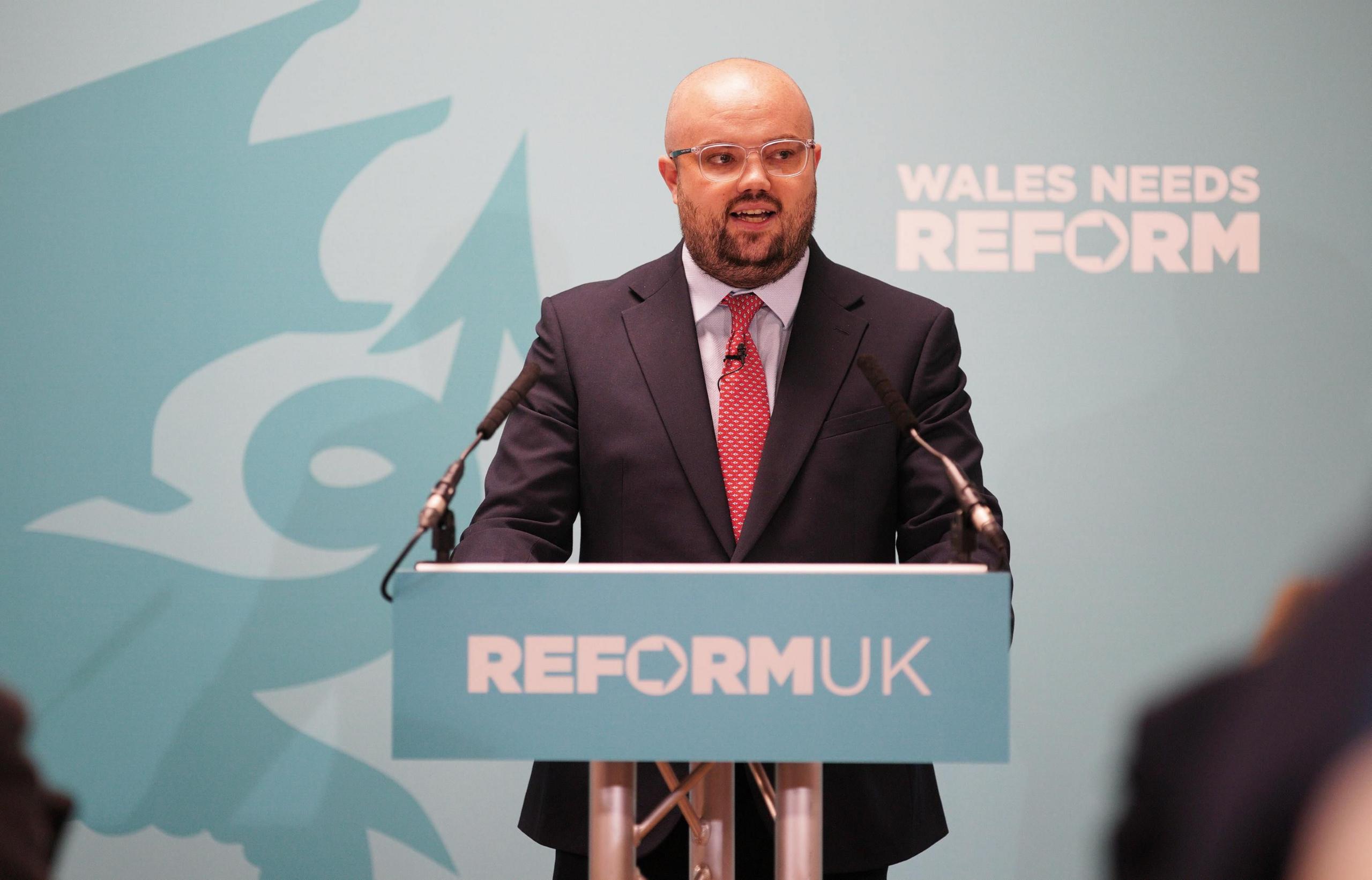 Man in blue suit, with a red tie, speaks at a podium.