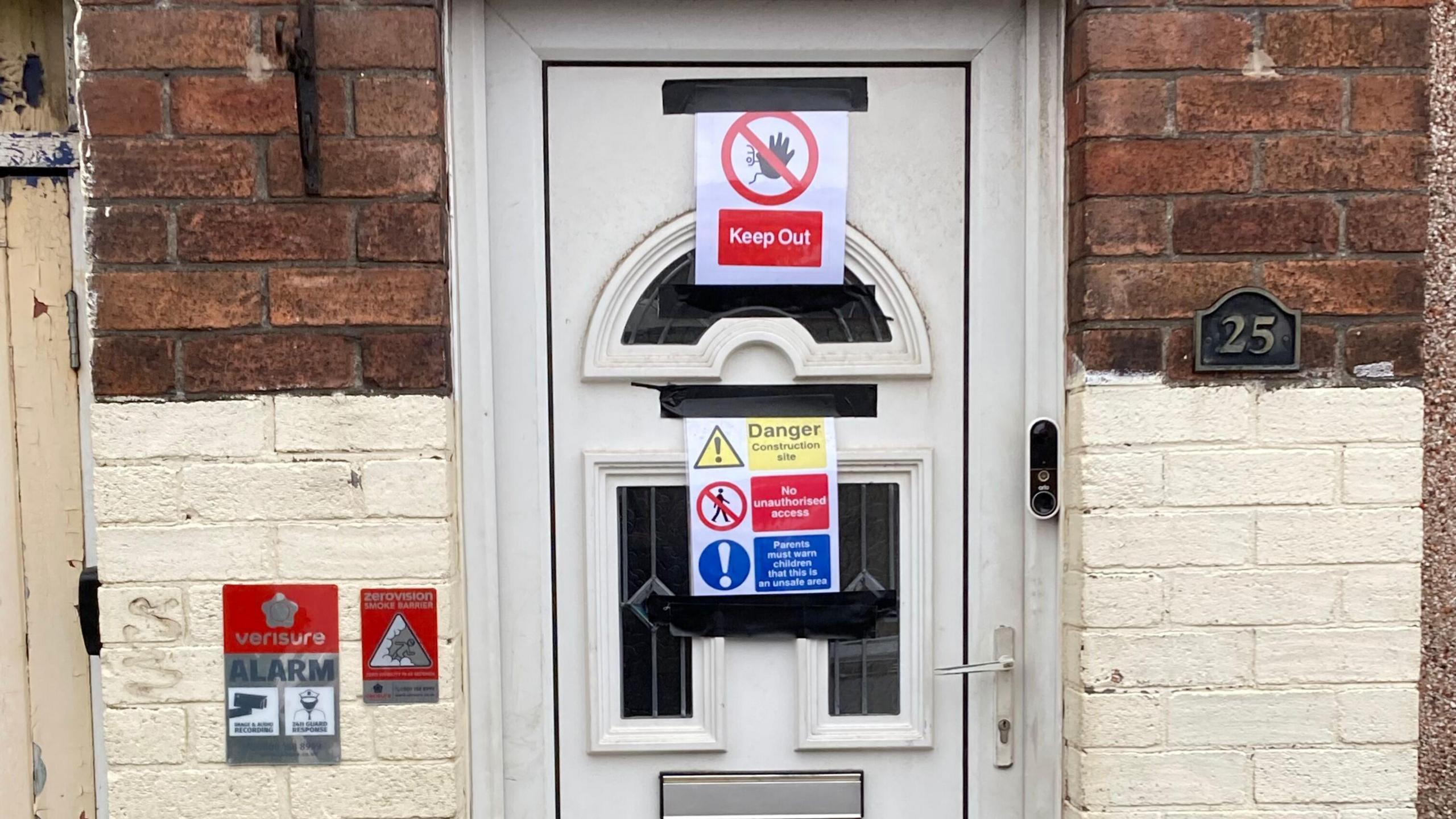 A white front door on a terraced property which has been affixed with warning signs such as 'Keep Out' and 'Danger, Construction Site'