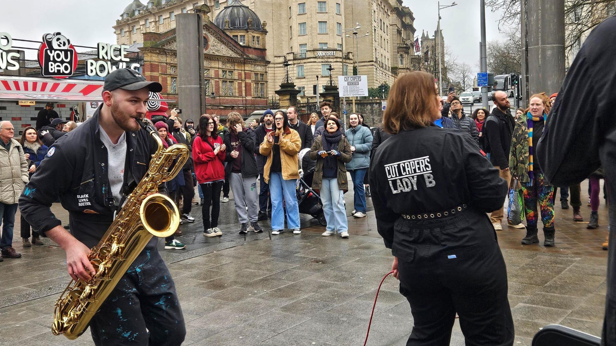 Two musicians from the band Cut Capers perform to a crowd of onlookers in the centre of Bristol, close to the harbour. The musicians are in black boiler suits with the name of the band on them, and many of the crowd are taking pictures