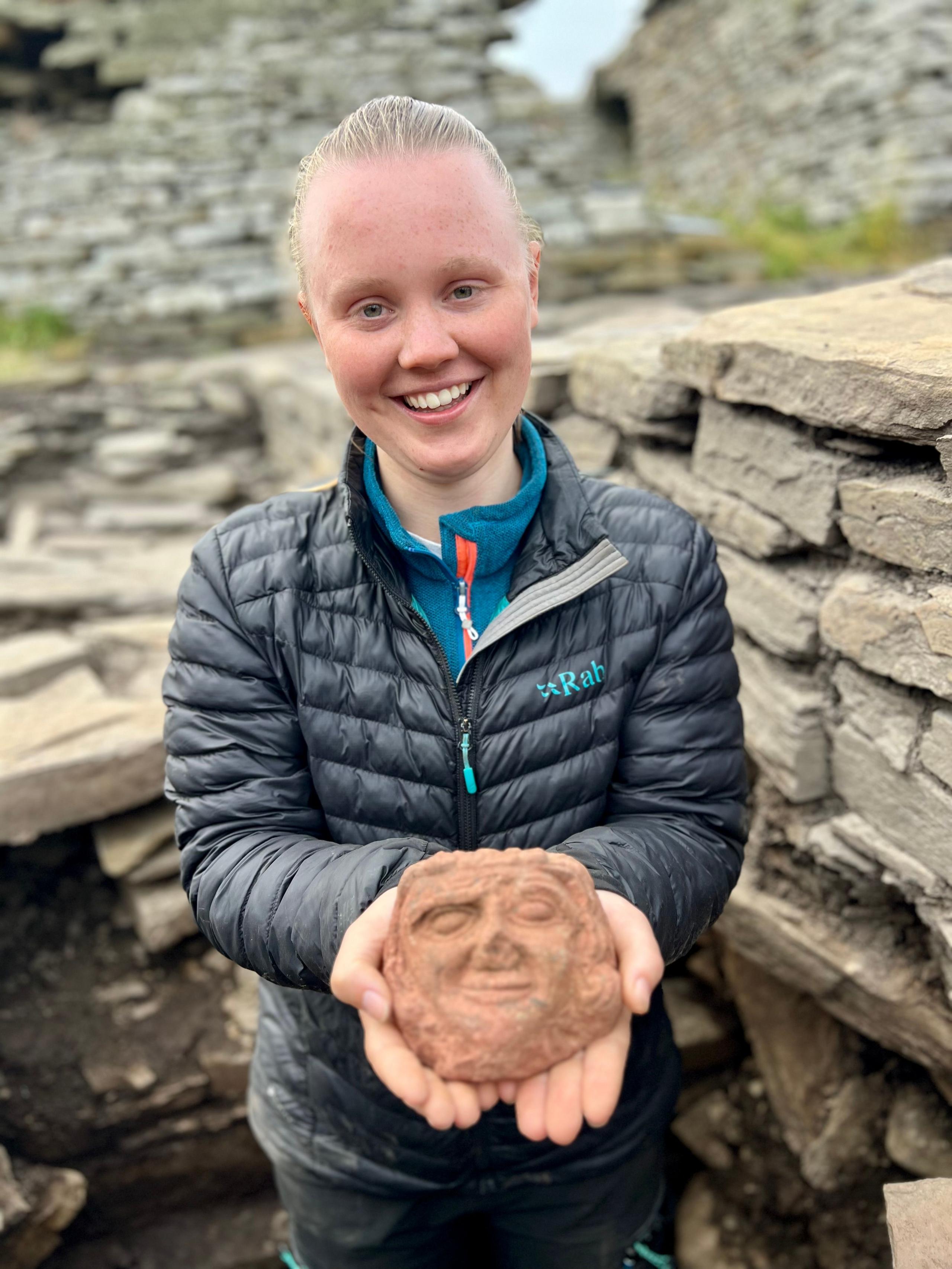 A young woman with hair drawn back in a dark padded jacket holds a sandstone carved face in her hands