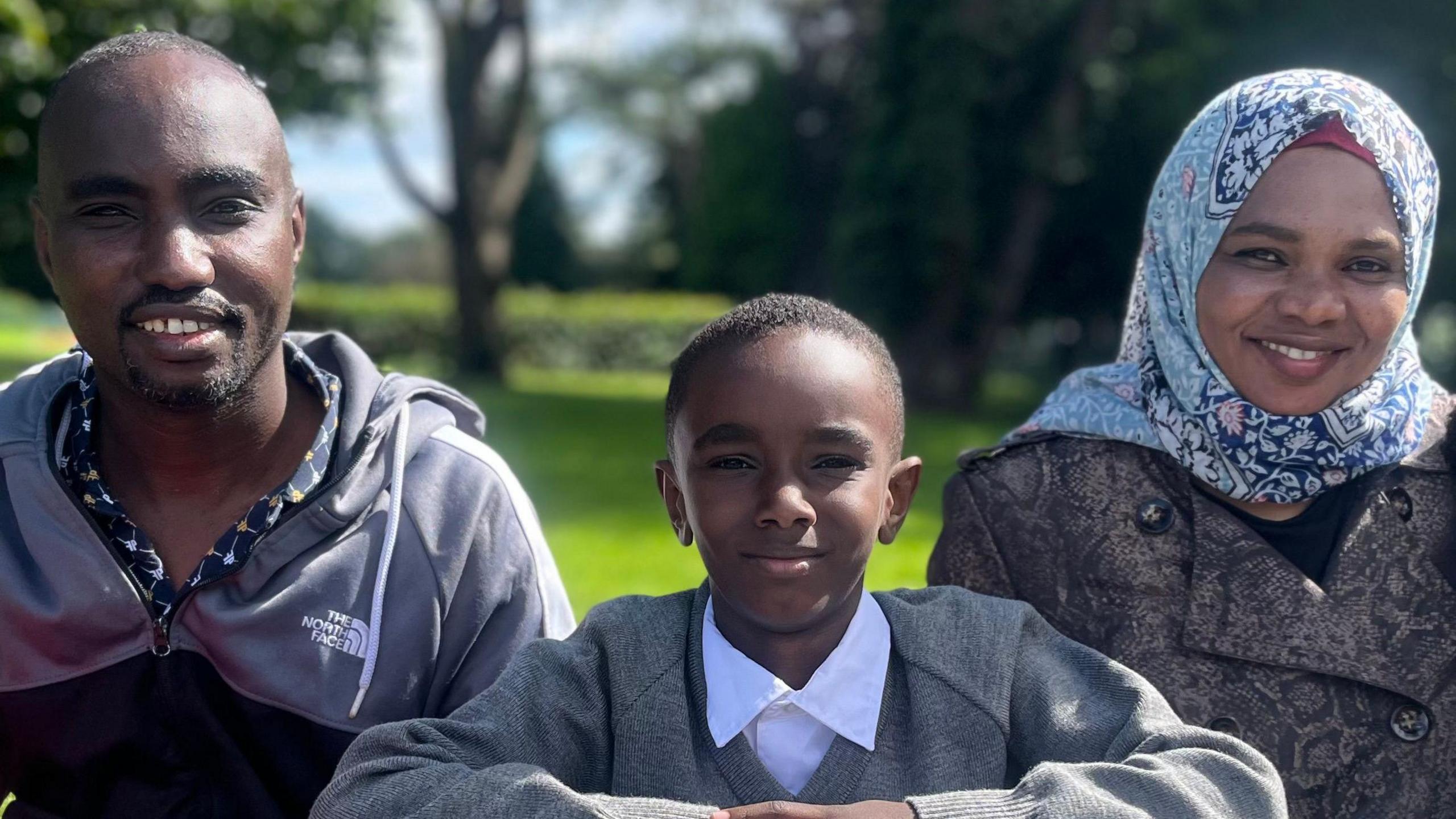 A boy in school uniform sitting in between two adults - a man in a shirt and hoody and a woman in a coat and a patterned headscarf. All three are smiling at the camera. They are in a green space and the sun is shining