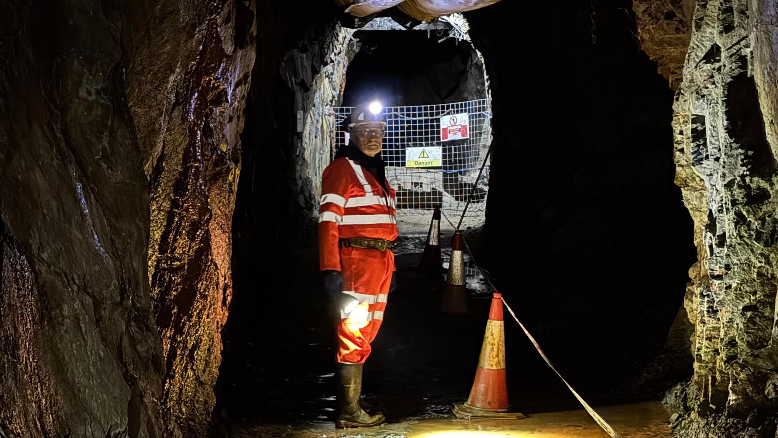 A man in hi-visability clothing stands in a mine shaft. He is wearing a hard hat with a head torch. He is looking back towards the camera, framed by rock walls, with the shaft continuing on behind him up to a gate that has fenced it off. 