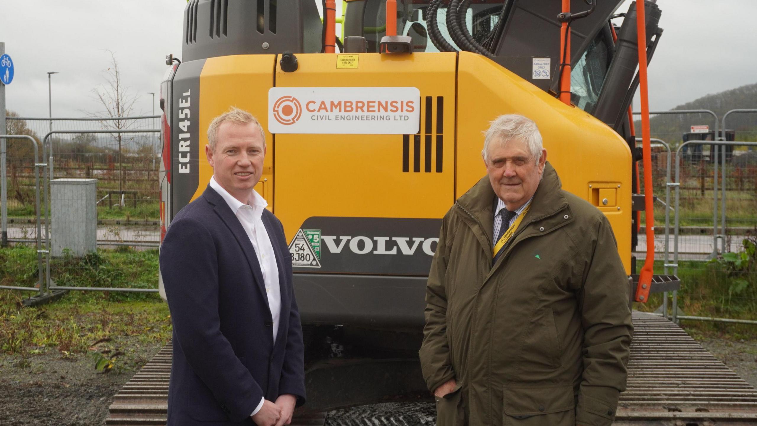 A man in a navy blue suit and white shirt stands beside a man in a dark green coat. In the background is a yellow Volvo digger and metal fences.