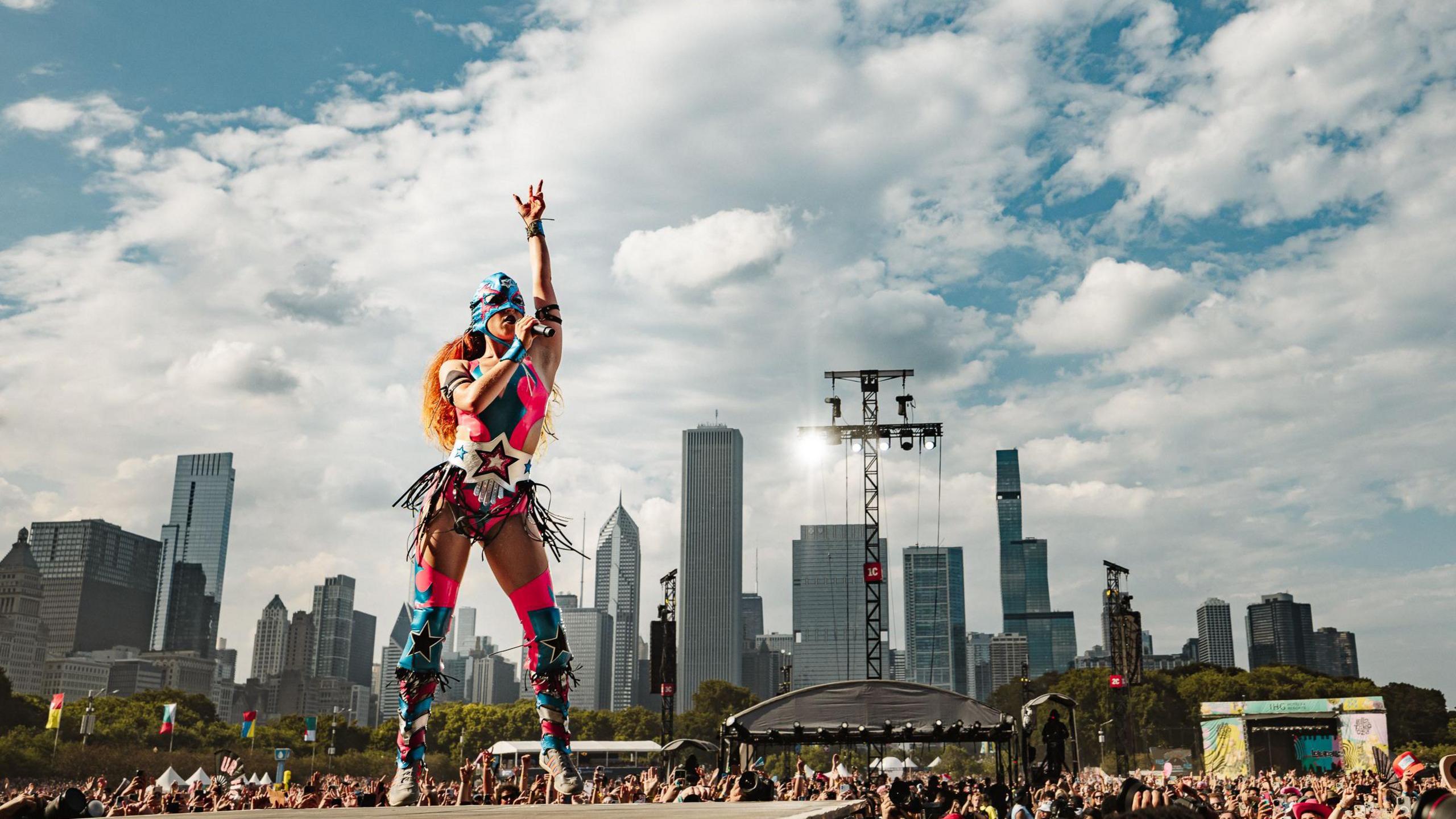Chappell Roan wears a pink leotard emblazoned with stars, thigh-high boots with stars and a super hero mask. She is standing on a stage with one hand raised in the air.