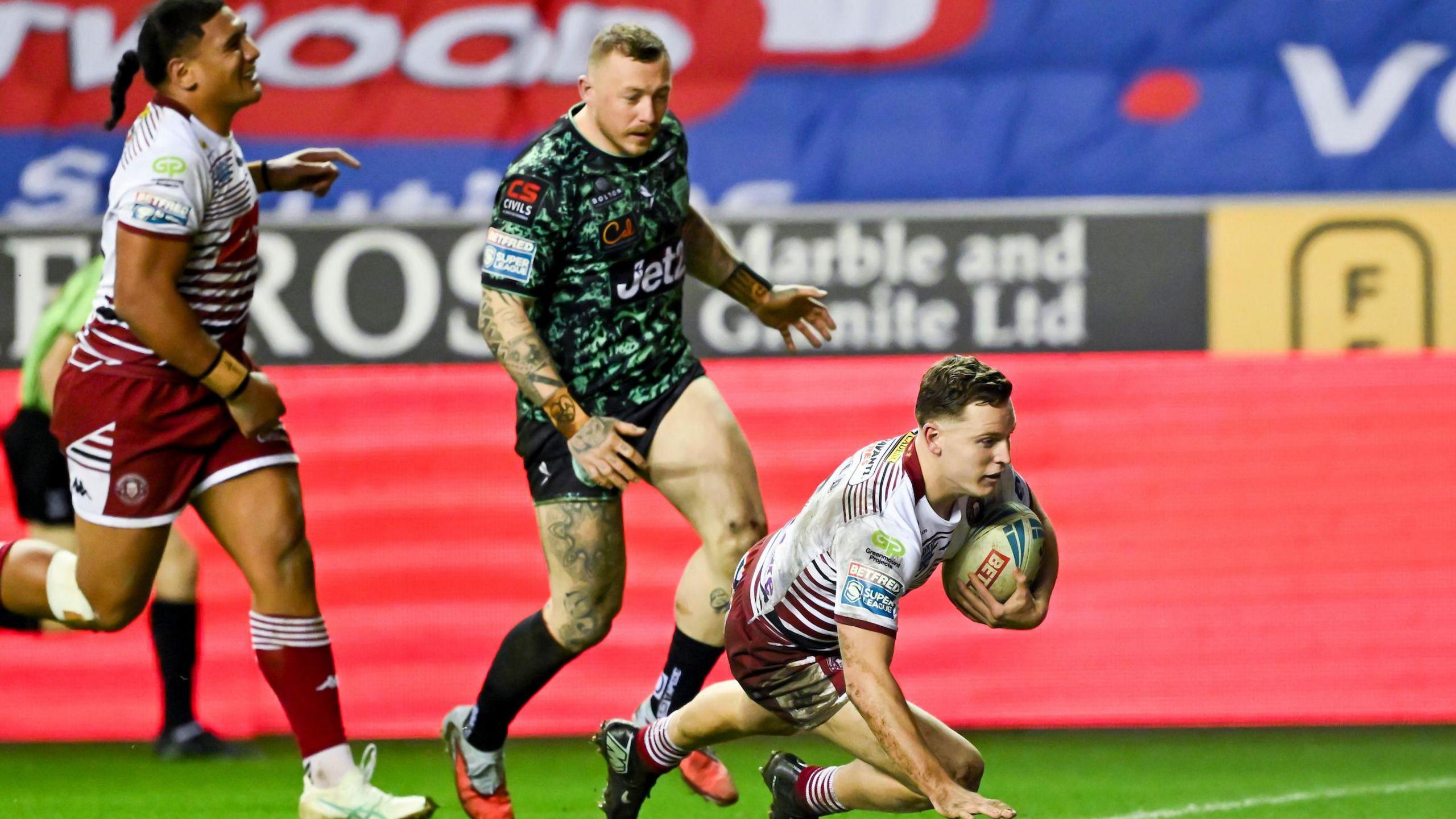 Jai Field, on one knee, about to touch down for a try with Leigh's Josh Charnley and Wigan team-mate Patrick Mago running behind him 