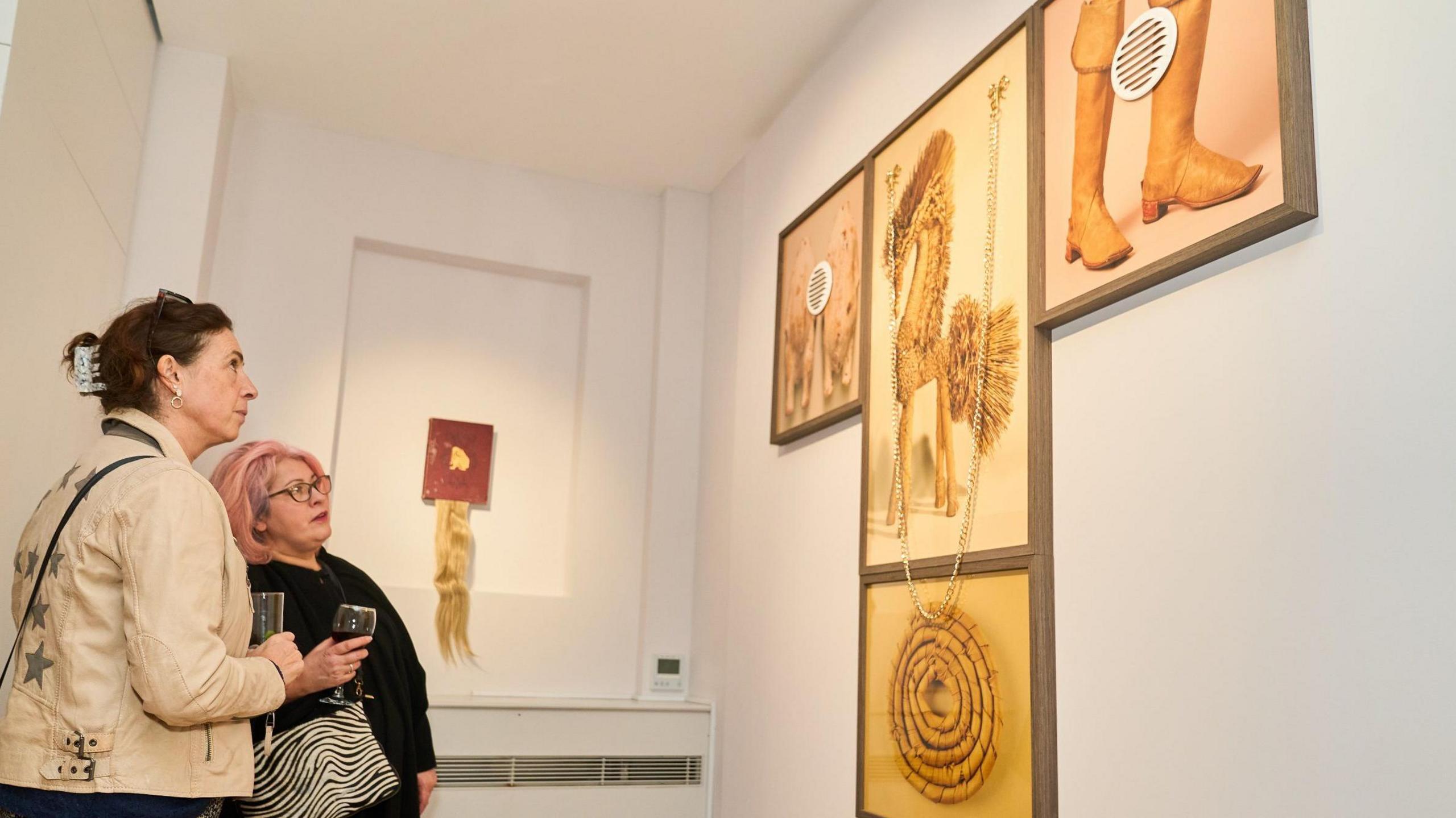 Two women holding a glass of wine each look at artwork on a white wall to the right of the image. There are framed photos of the following, all in a sepia brown colour: a coiled rope, a pair of brown cowboy style boots, a strange creature like a seahorse-horse hybrid, and two dogs. On the other wall is a wig of blonde hair exhibited between the pages of a book.