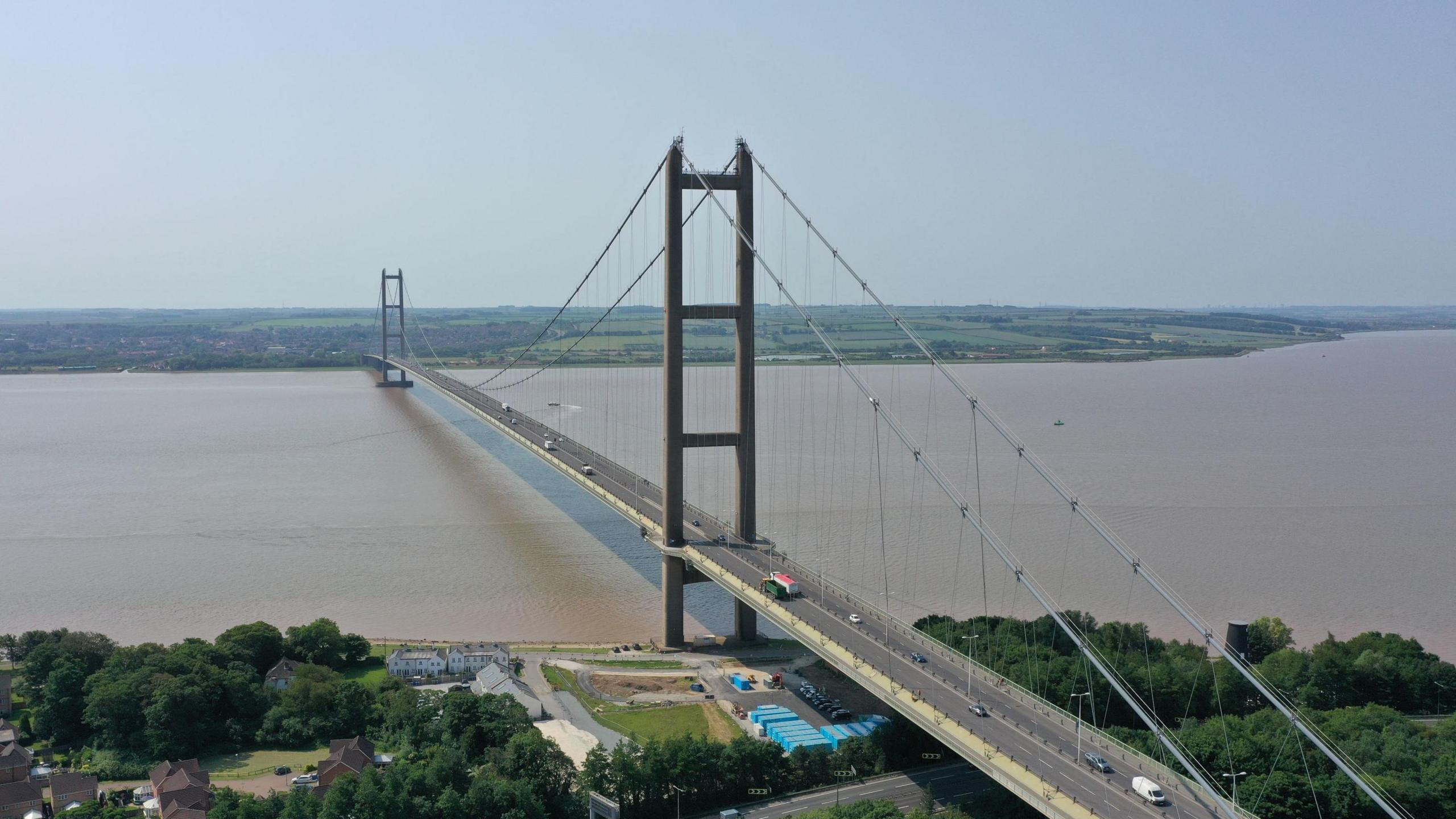 An aerial shot of a large bridge spanning a wide brown river, with tall towers and cables holding it up. Cars are travelling across the bridge. There are houses, trees and a small construction site near the bridge's base in the foreground of the photo.