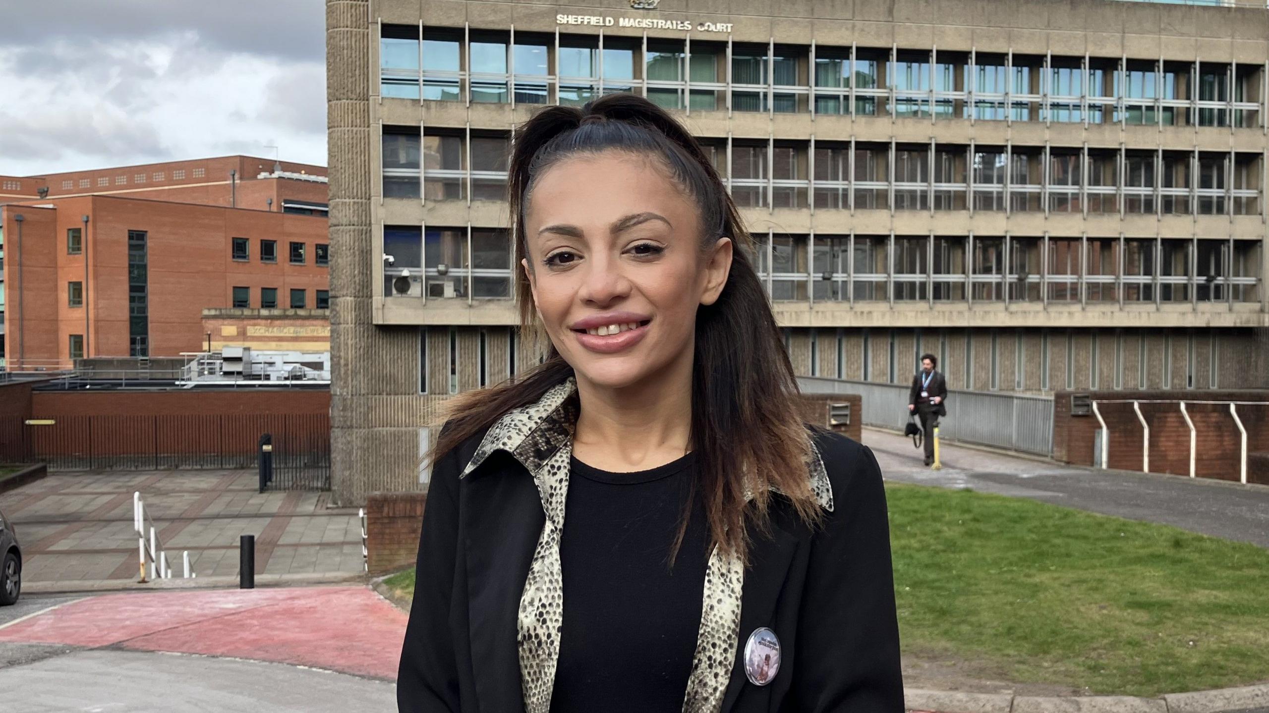 Sophie Zaherali smiling outside Sheffield Magistrates' Court after her hearing. She is wearing a black blazer and a snake-print shirt over a black T-shirt. Her dark hair is in a high ponytail.