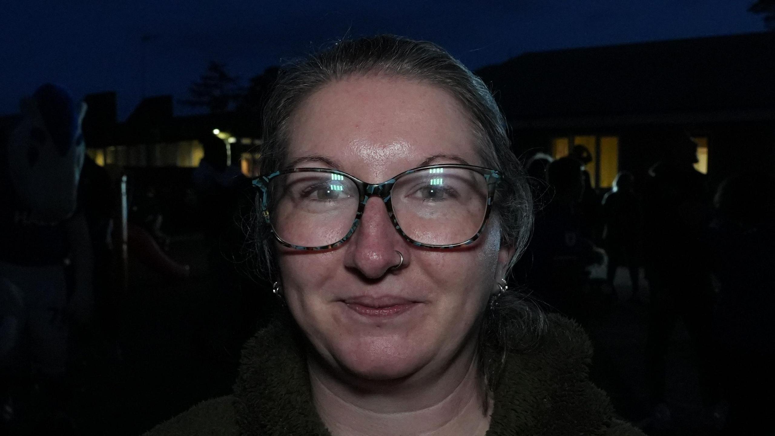Laura-Jane Elmer smiles at the camera as she stands outside a family hub at night. She has grey hair that has been tied up behind her head and she wears glasses and a green fluffy jumper.