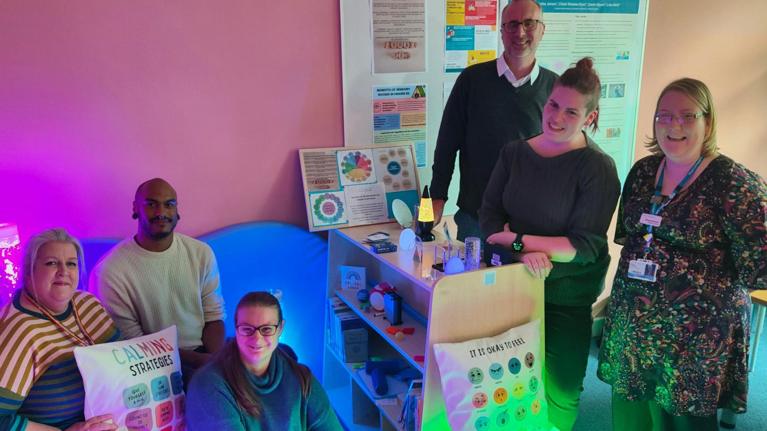The picture shows a small sensory study space with calming features. There is a shelving unit holding items such as fidget toys, small containers, and books. The room is softly lit with coloured lights, including purple and green tones. Behind the shelving unit, there are posters on the wall with diagrams and information.