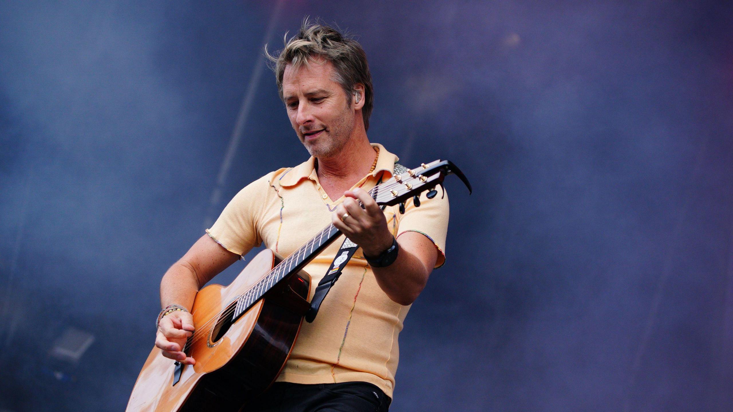 Chesney Hawke on stage with an acoustic guitar - he is wearing a yellow polo shirt