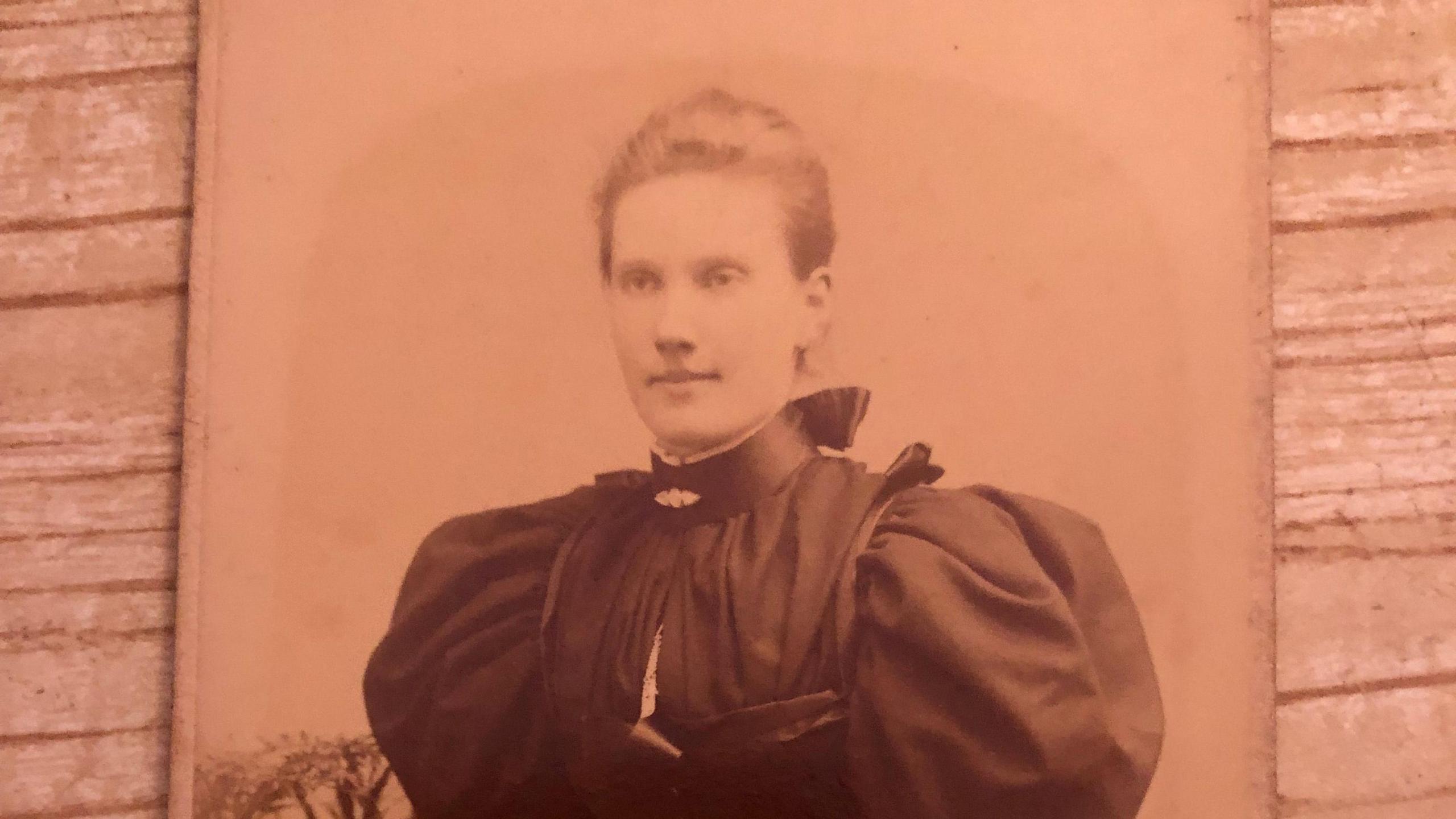 A Victorian sepia photograph of Ann Walton. She has light brown hair pulled back into a bun. She has a dark high-collared dress with puffed sleeves with a bodice and a white shirt underneath.