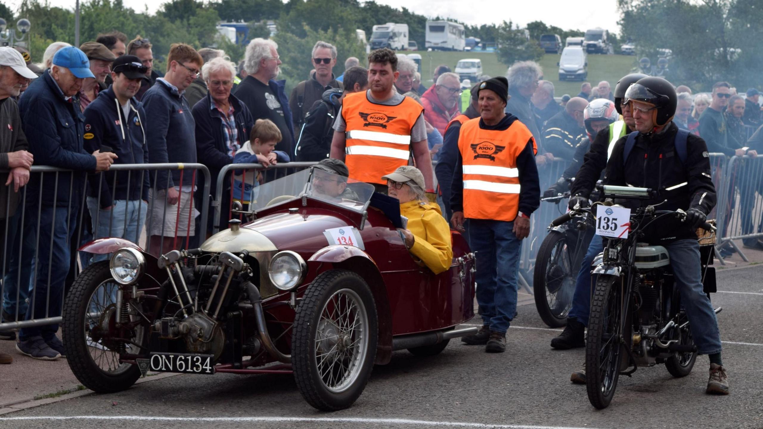 A man and a woman sit in a vintage two-seater car, next to a man on a motorcycle. Crowds of people are watching from the other side of a barrier.