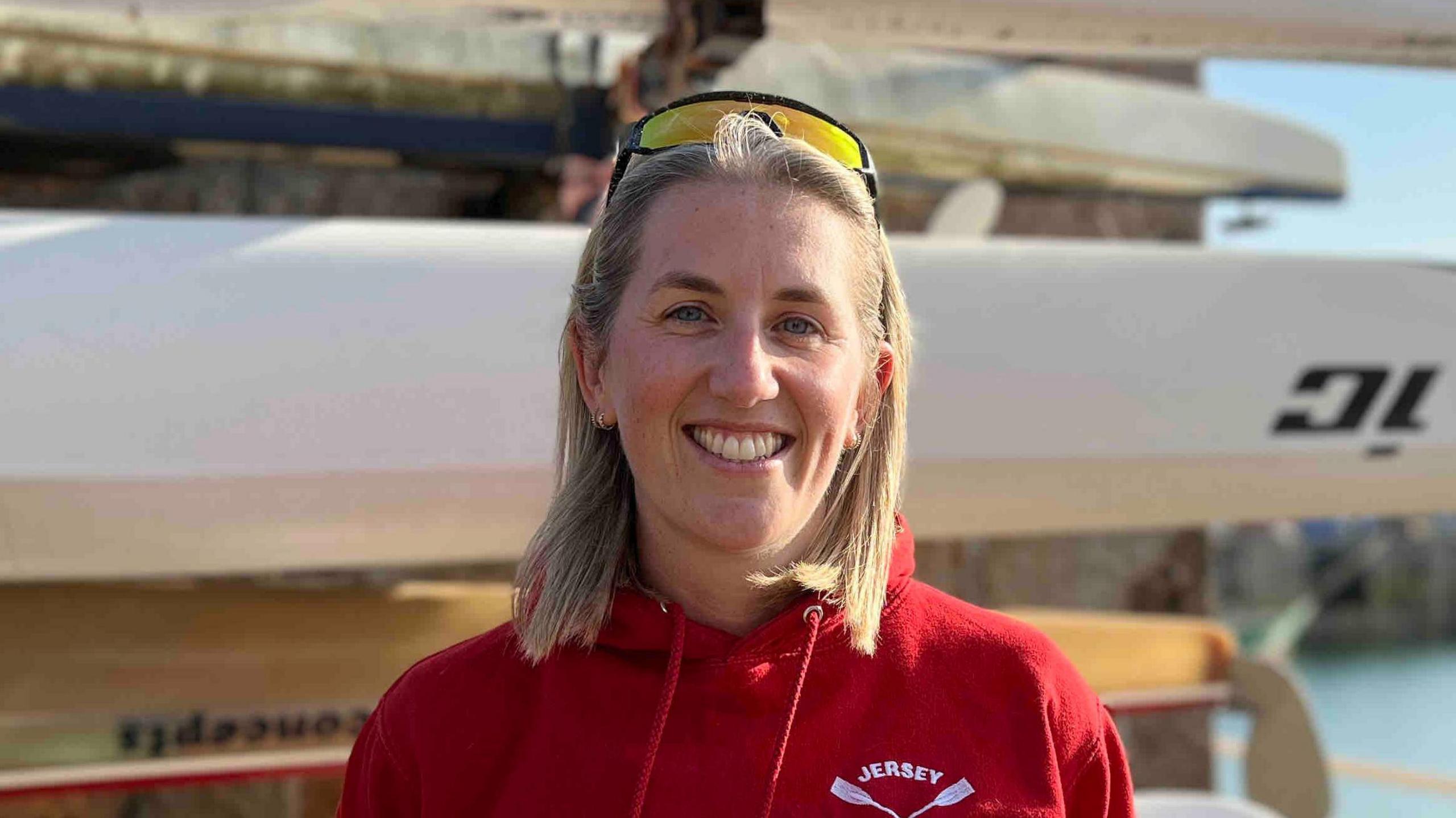 Emma is wearing a red 'Jersey Rowing Club' hoodie standing in front of stacked kayaks by a waterfront, with calm water, stone wall, and distant buildings under a sunny sky.