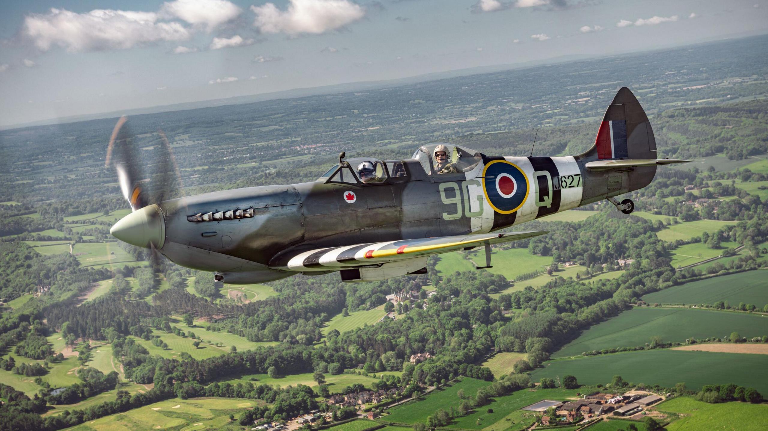 A Spitfire flying above green fields and trees. it has the Royal Airforce emblem on the side.
