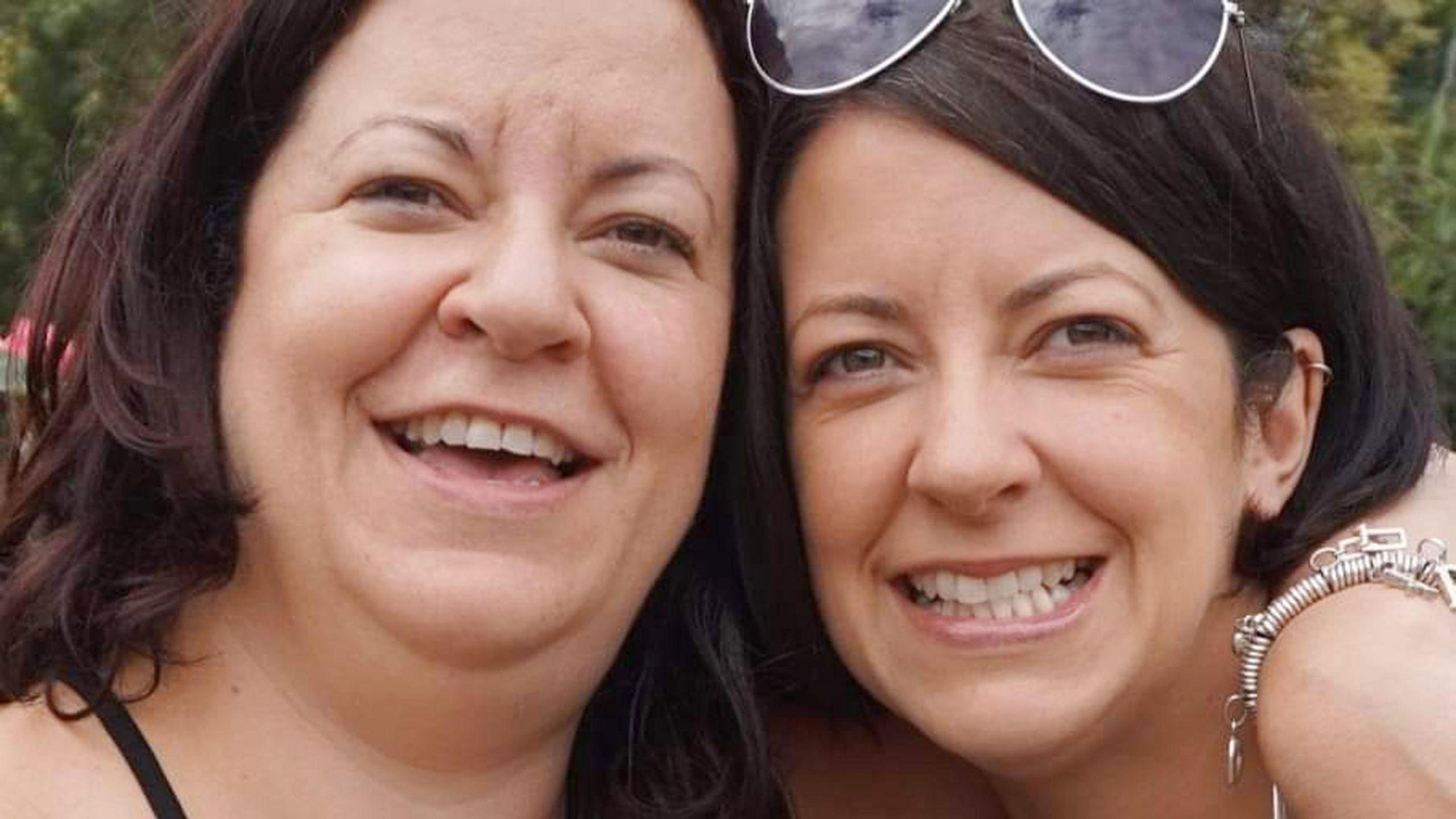 Emma and her sister Kelly have their arms around each other and are smiling. Kelly, on the left, has red brown hair. Emma has brown hair and a pair of sunglasses on her head