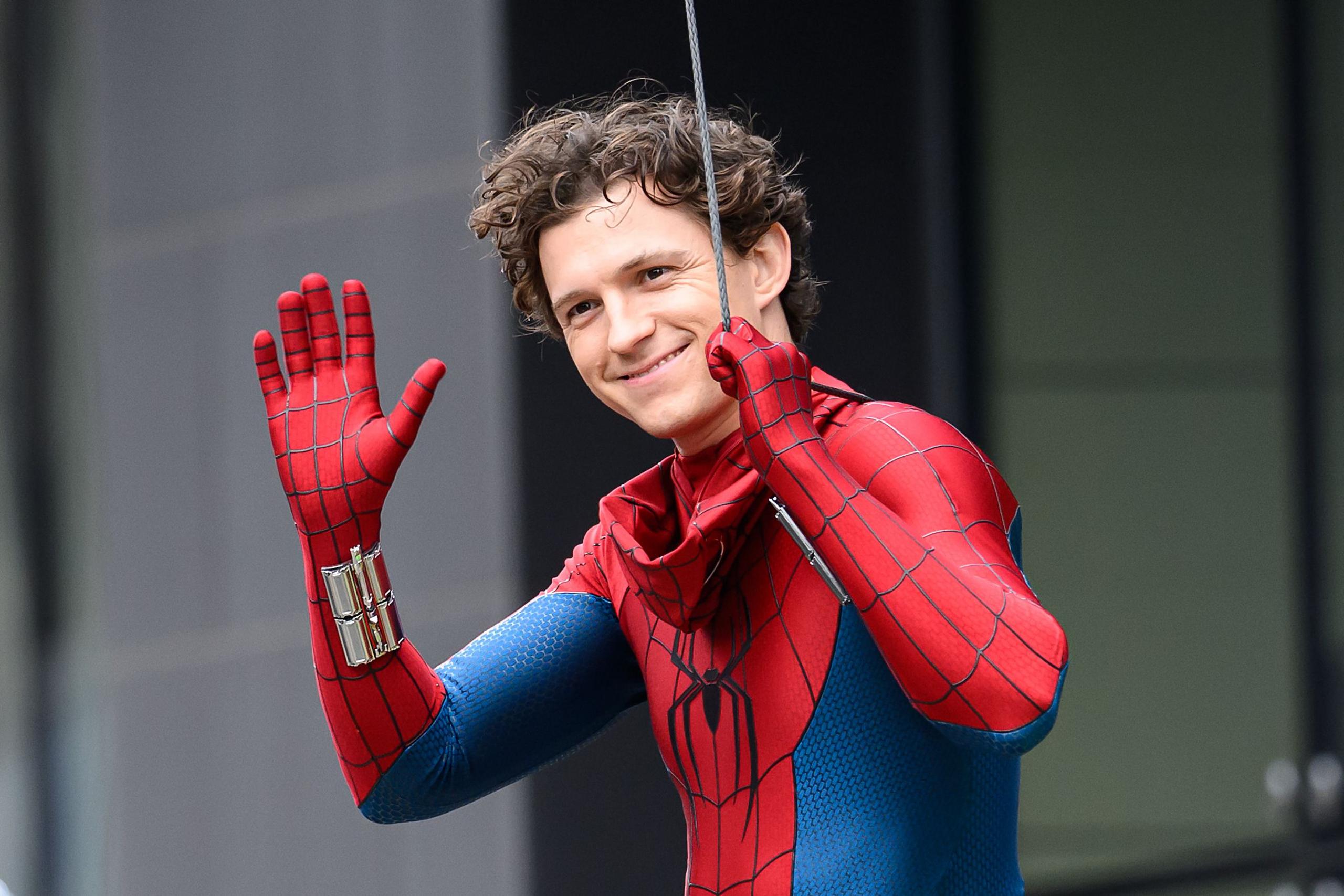 Tom Holland smiling and waving to fans while in costume and holding on to a thick wire which is attached to his back.
