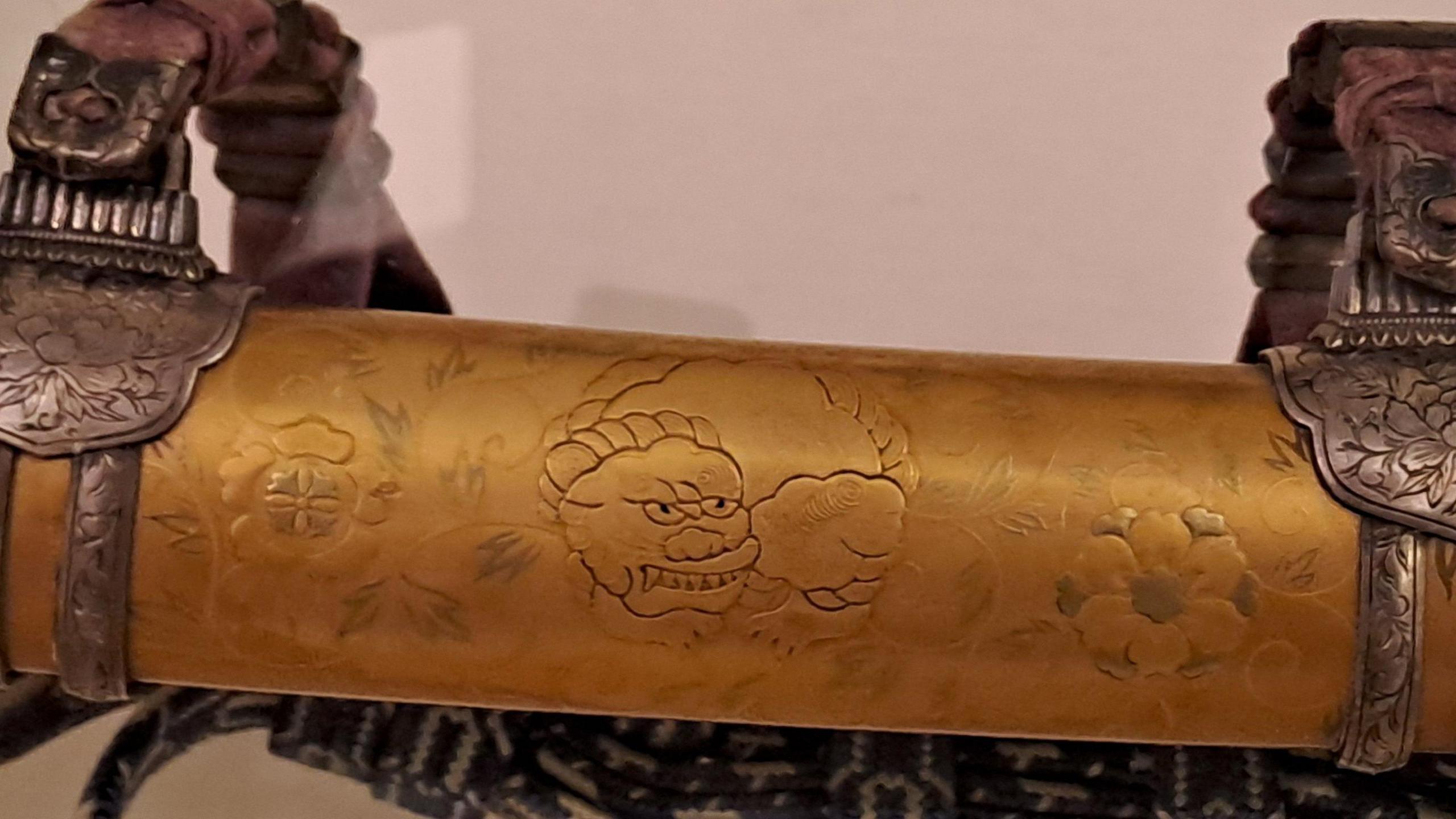 Close-up of an ornate sword mount featuring a golden scabbard with intricate engraved floral patterns and a stylized lion-like face motif. The mount includes two decorative metal fittings with embossed floral designs and attached leather straps.