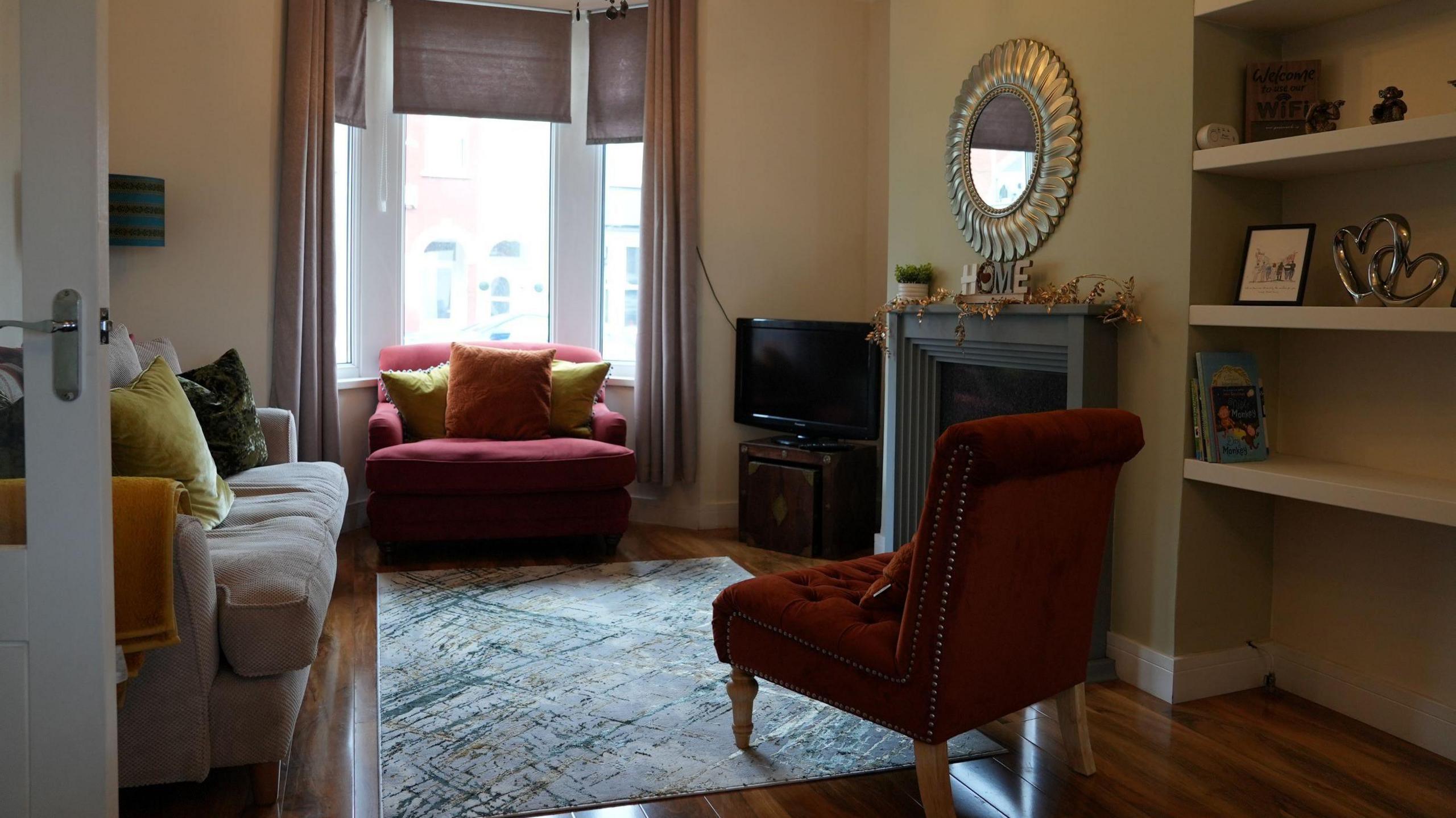 Photograph of the main living room on Trinity Street, Barry, Vale of Glamorgan. There is an open fireplace on the wall on the right hand side, and a grey sofa on the left. An orange armchair takes up the bay window, covered in mustard and orange velvet cushions. A velvet orange chair edges a grey rug in the centre of the room. The walls are a very pale green, a TV stand is in the corner of the room, and shelves can be seen on the right hand side of the fireplace. A round mirror is on the wall above the fireplace. 