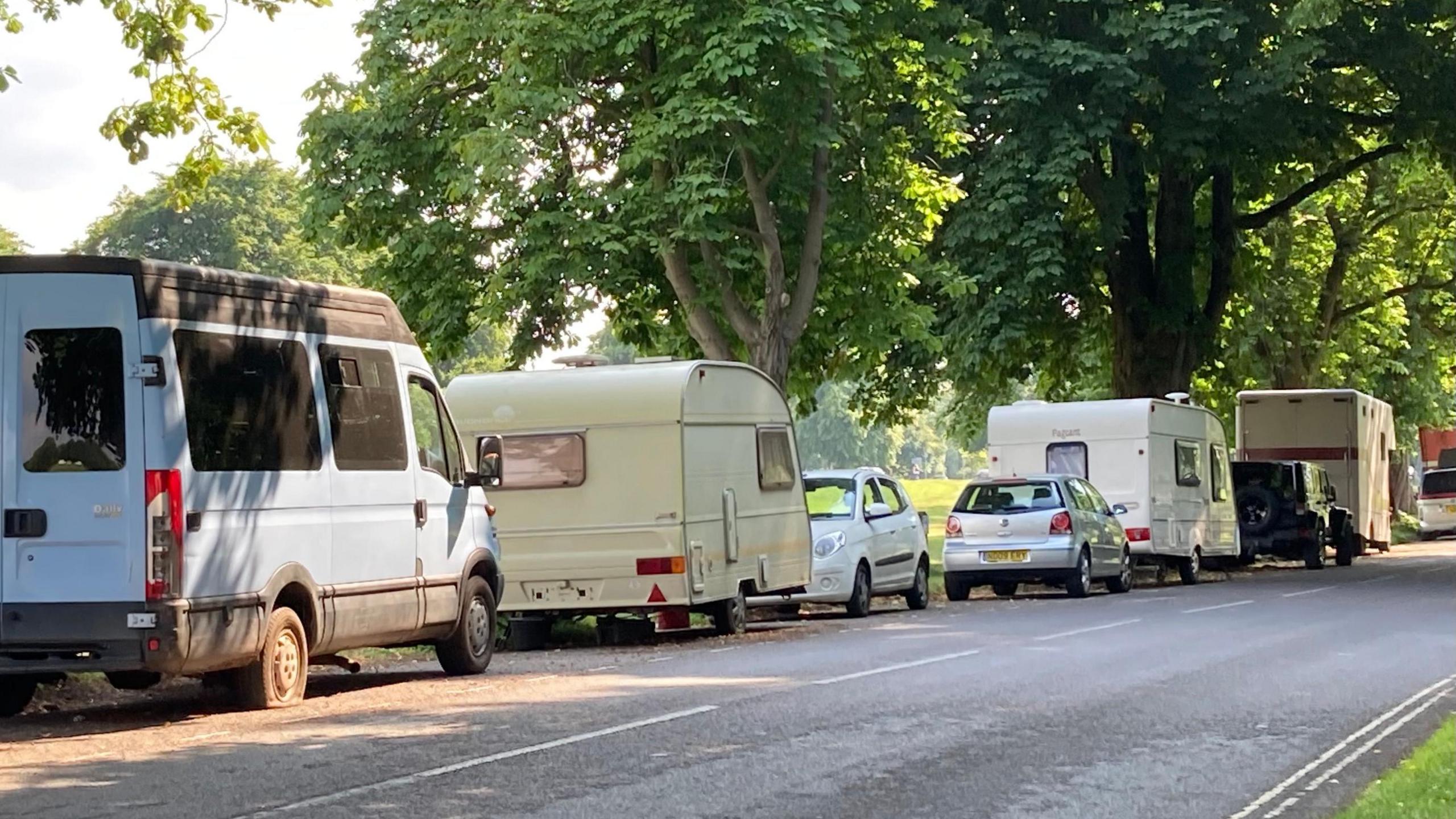 A line of white vans and caravans, some with cars in front or behind, parked along a section of road along The Downs in Bristol