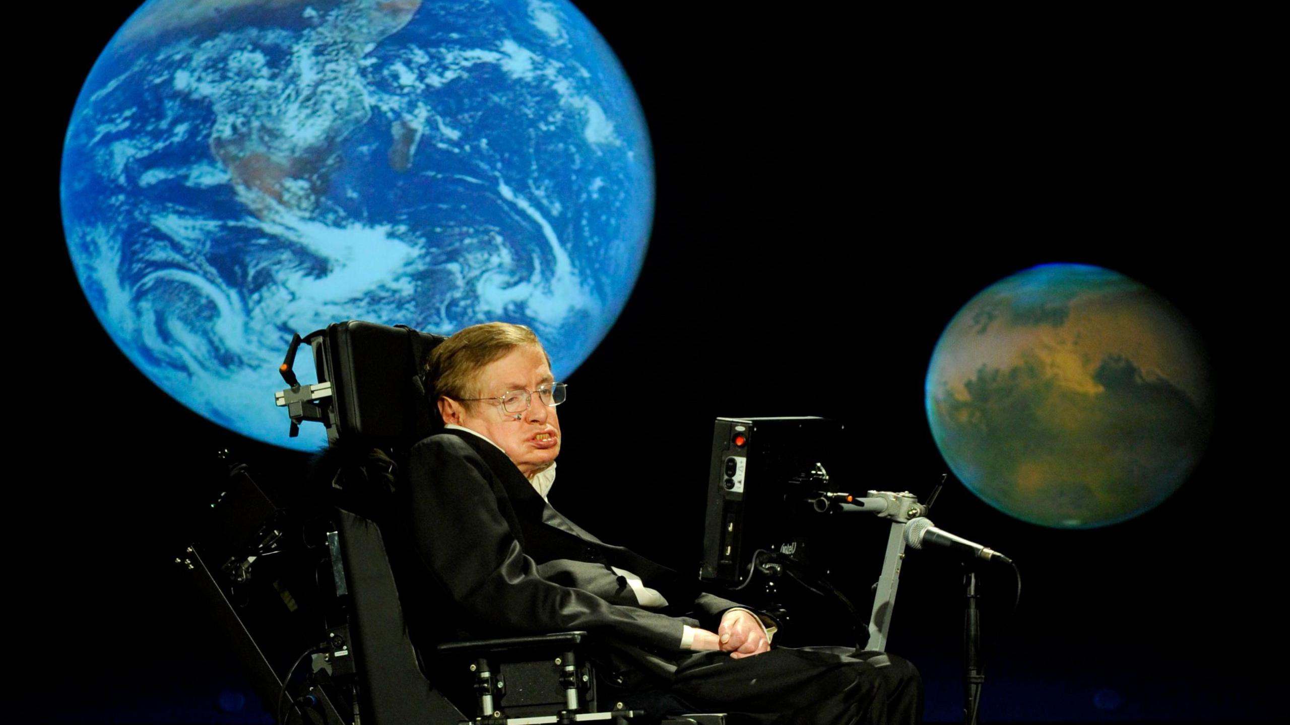 Stephen Hawking on stage giving a lecture in front of an image of planet Earth.