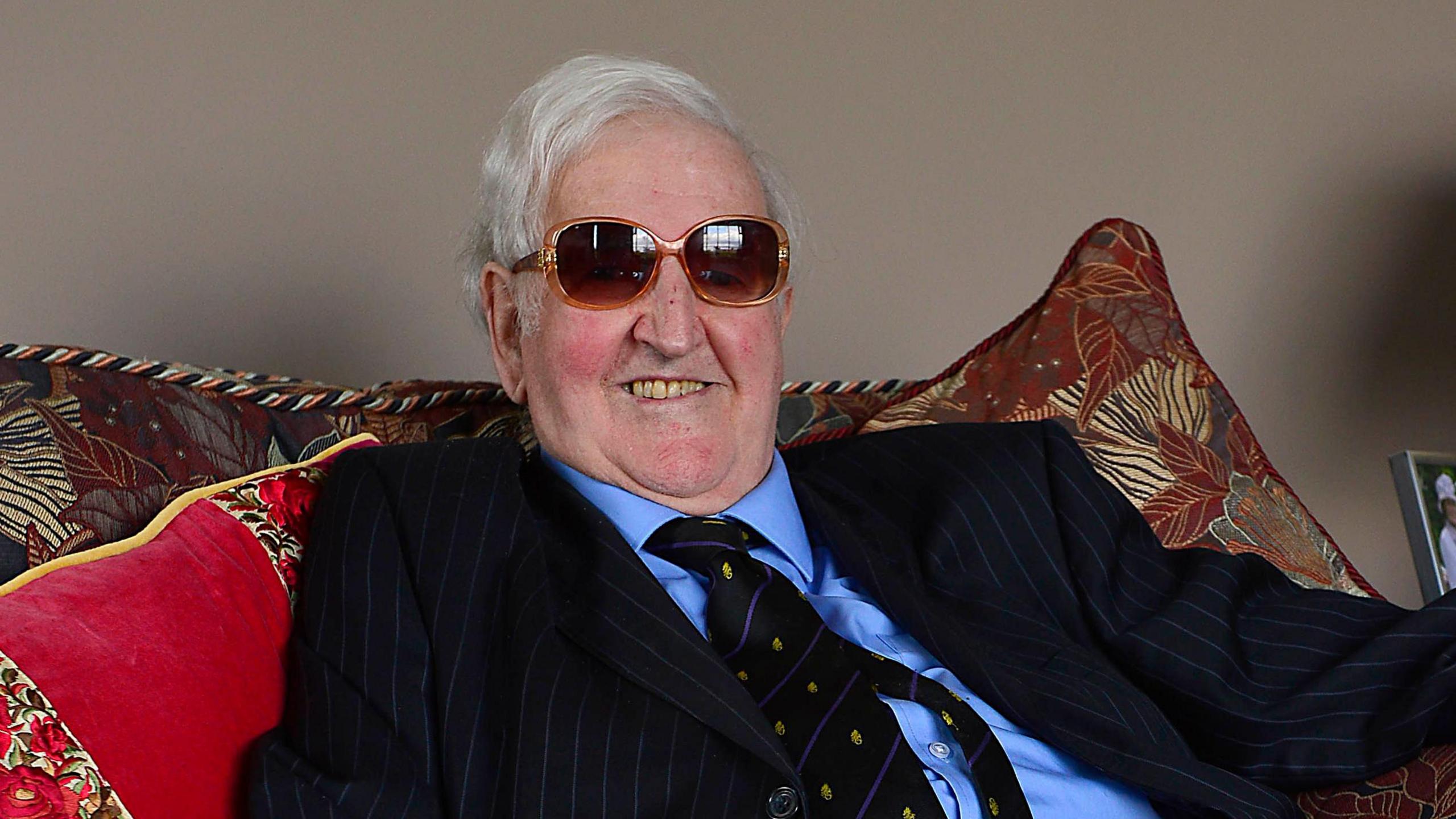 Sir Kenneth Bloomfield has grey hair and is wearing brown sunglasses. He is sitting on a red and brown sofa and is smiling at the camera. He has a navy suit on with a blue shirt and striped tie.