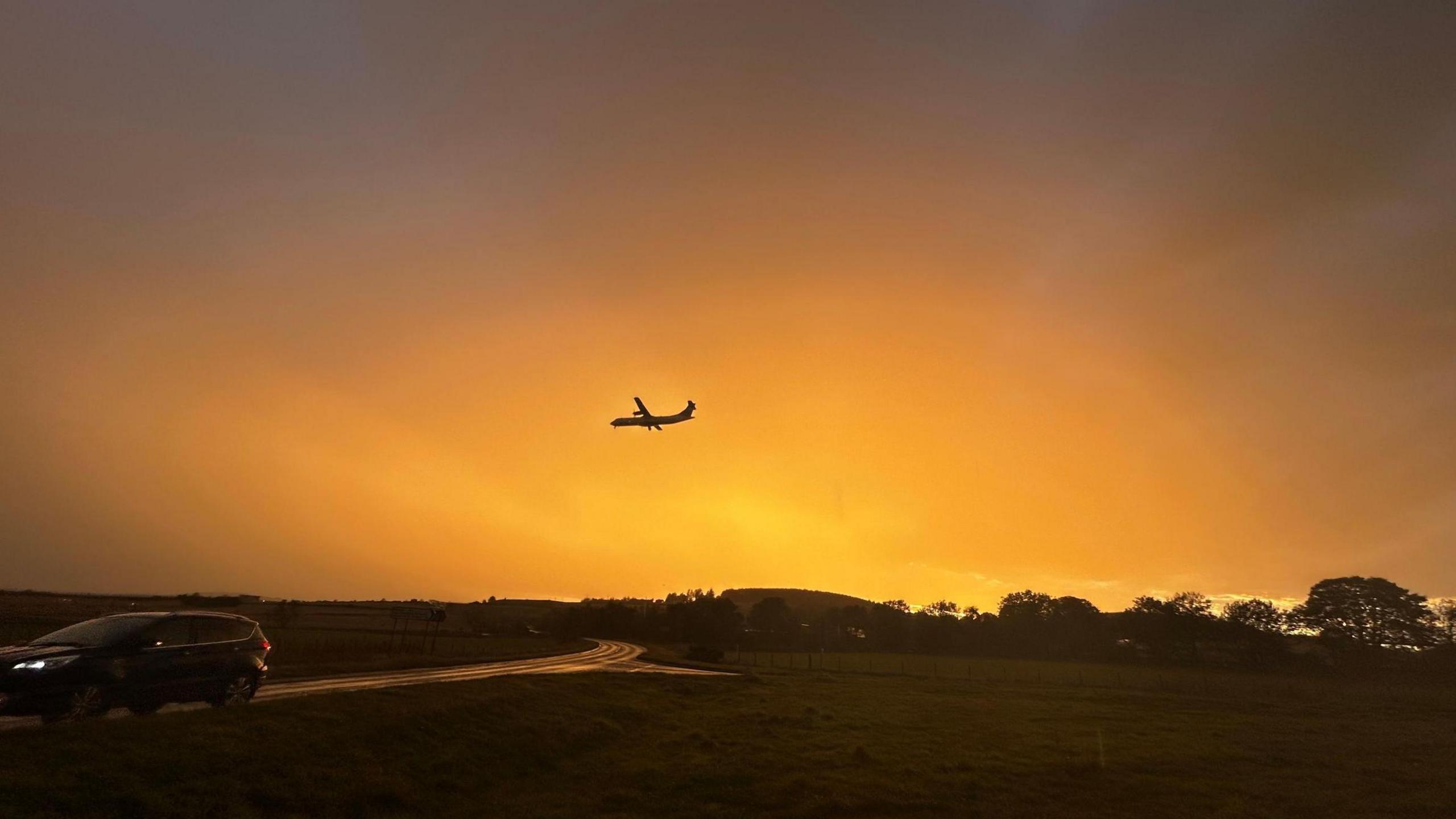 An orange evening sky at Oldmeldrum in Aberdeen. A passenger plane can be seen coming into land at Aberdeen Airport, which is out of view, while a car passes on a road in the foreground.