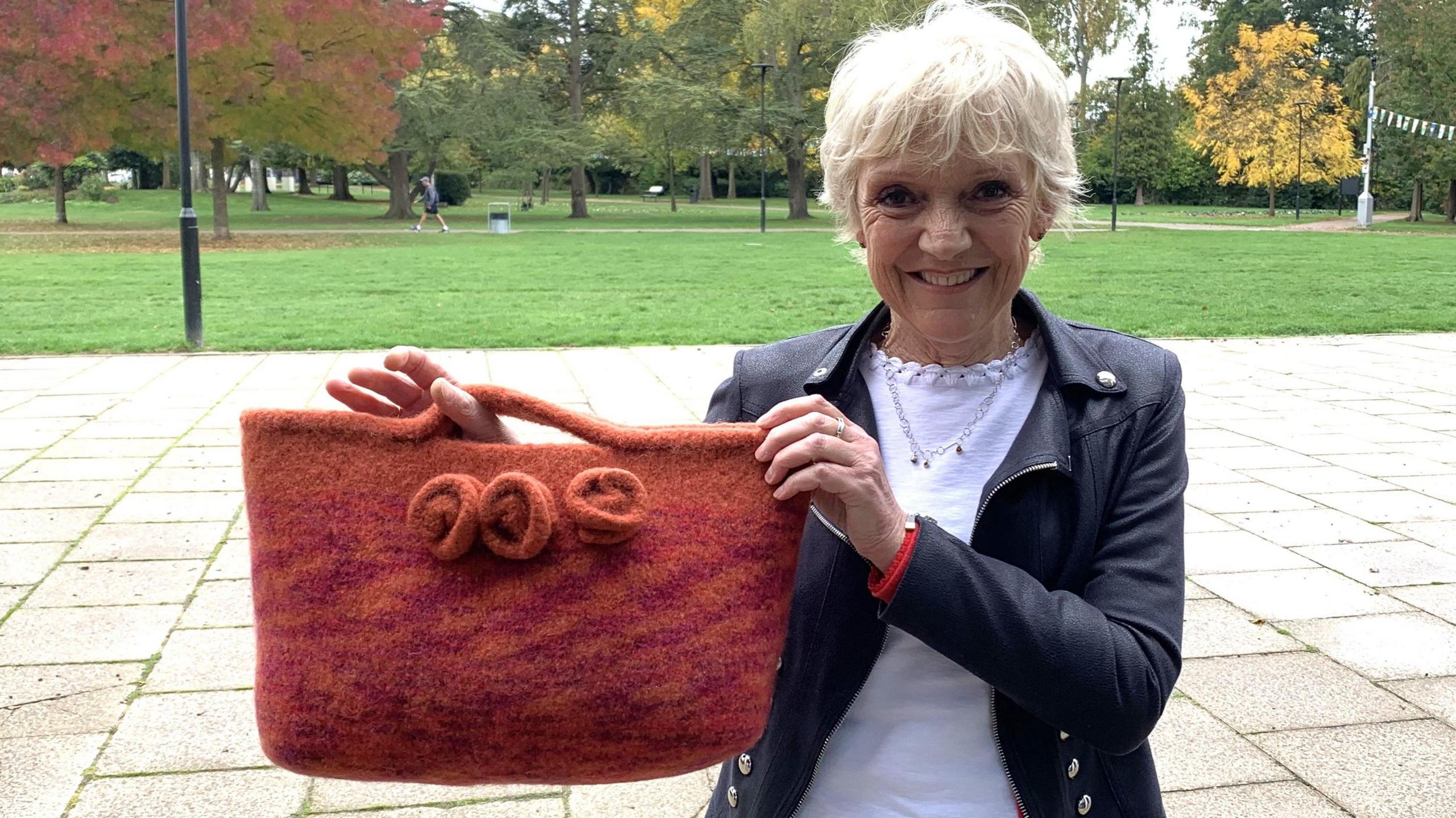 Gillian Wright stands on paving with a park and autumn trees behind her. She holds up a burnt orange crocheted bag with 3 roses on the front. She has short blonde hair and wears a black leather jacket and white T-shirt